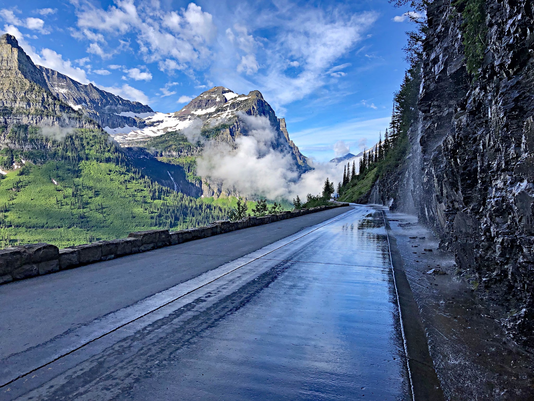 Cycling Logan Pass West, Going-to-the-Sun Road, Glacier National Park - wet portion of straight roadway, water runoff on wall alongside road, sharp, steep mountain peeks in distance shrouded in fog