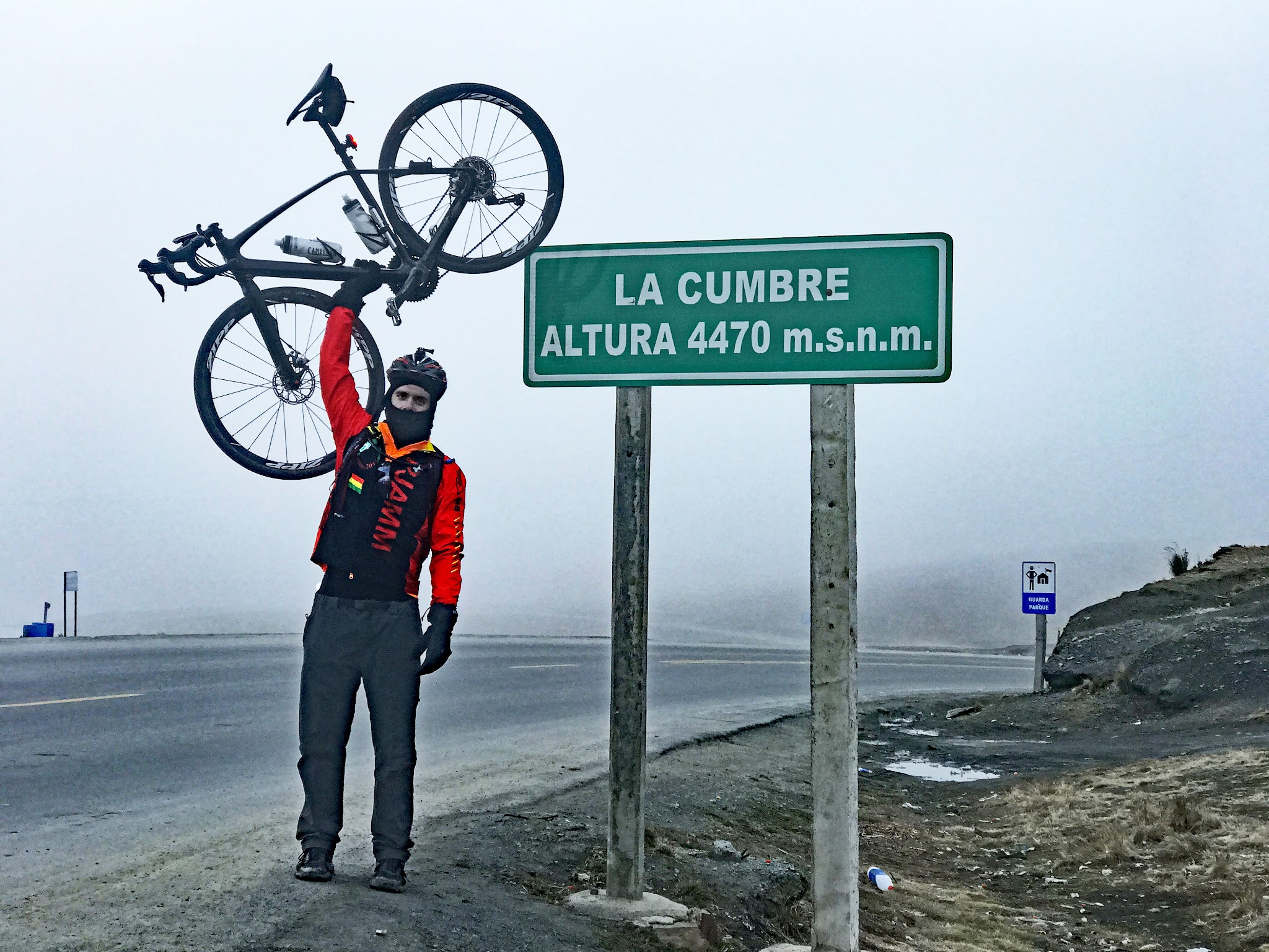 Cyclist raising bike at La Cumbre Peak summit and elevation sign