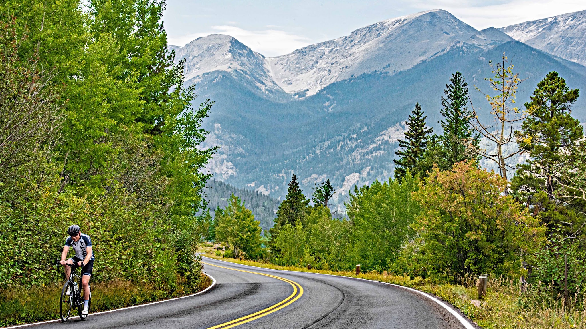High Point Cycling Trail Ridge, Colorado - cyclist climbing on two-lane roadway surrounded by greenery, mountains in background