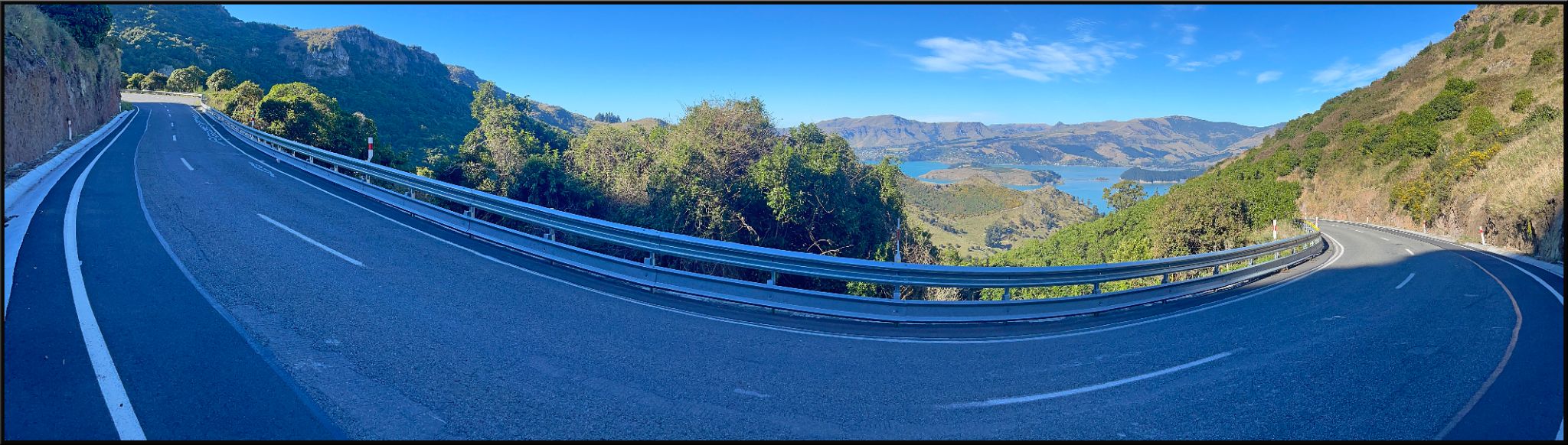 panoramic view of roadway with guardrail, overlooking mountain terrain with Governor’s Bay on Banks Peninsula in far distance