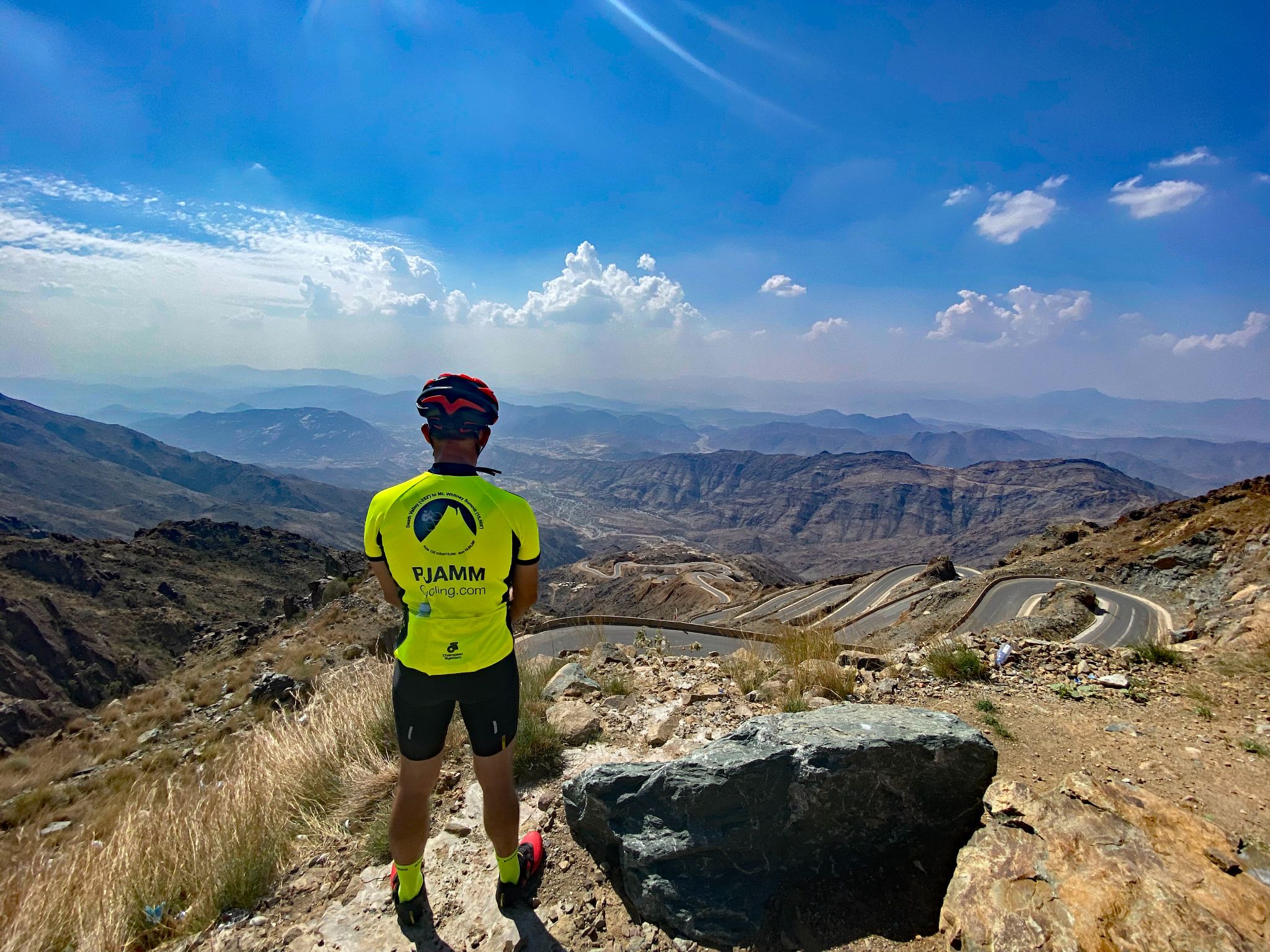 Cycling Bashout Pass, Saudi Arabia - PJAMM Cyclist stands next to a rock on the side of the roadway, looking down on the intense hairpins in the roadway below