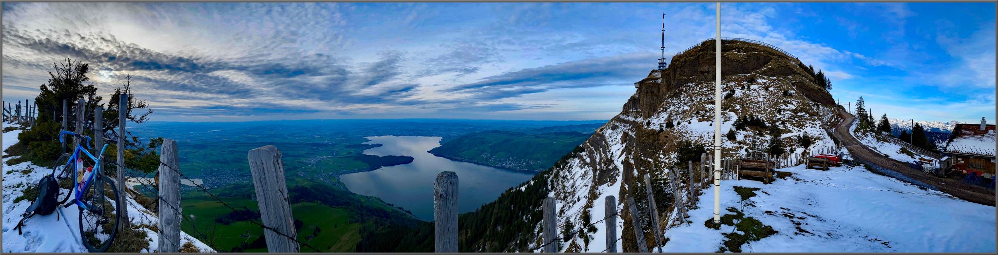 Panoramic views of snow capped mountaintops, bike leaning against fence on mountain side, lake in the distance