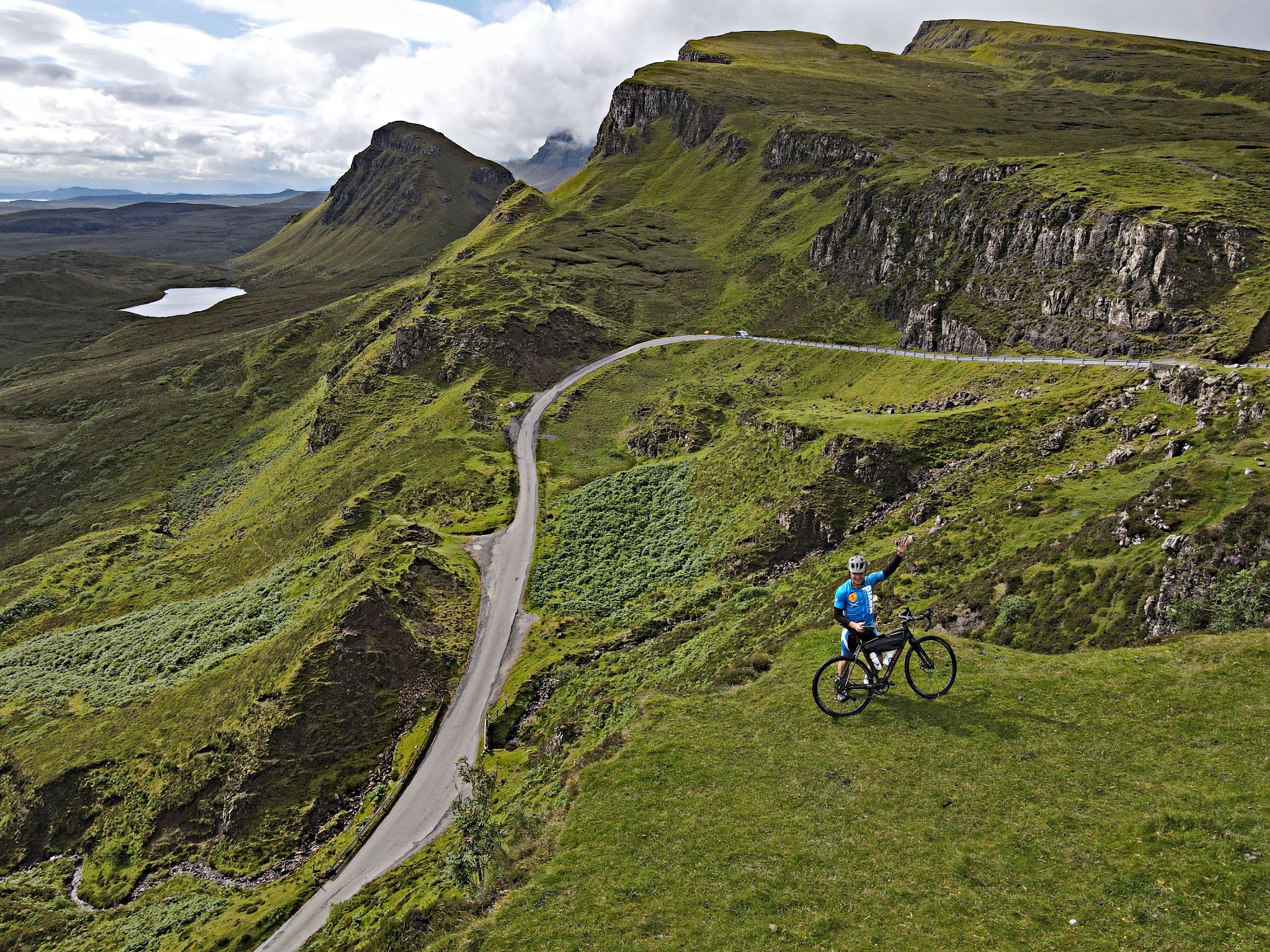 PJAMM Cyclist stands with bike on green cliffside, waving to aerial drone camera, roadway far below