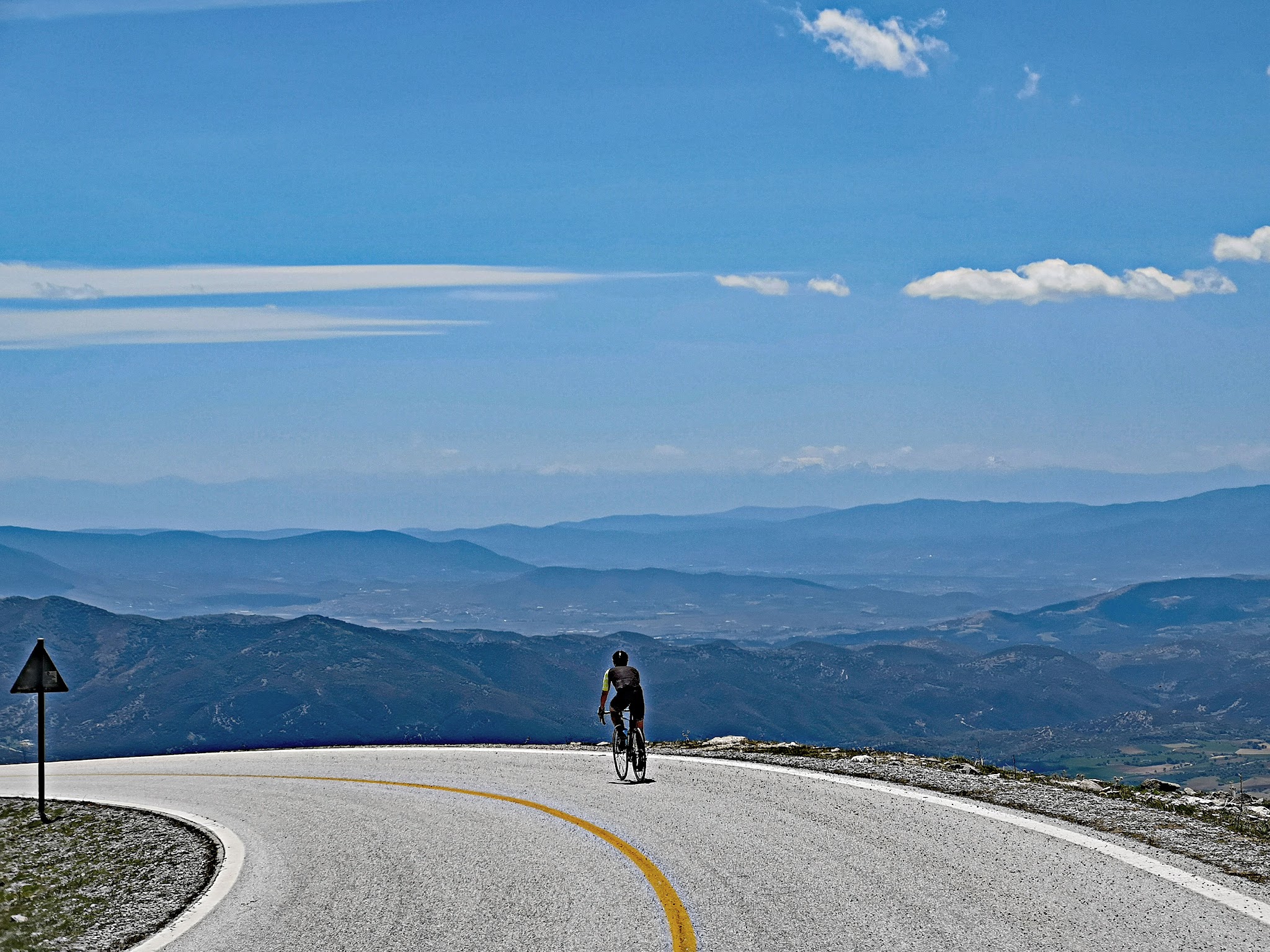 Cycling Mount Olympus, Greece - cyclist rides on curve in two-lane roadway on mountainside, mountains can be seen layering the horizon, white clouds in blue sky
