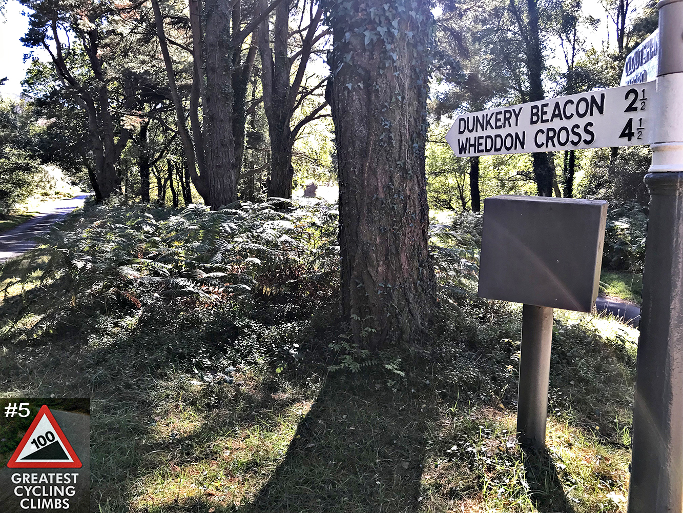 Cycling Dunkery Beacon, UK - road sign for Dunkery Beacon and Whedon Cross, large trees and ferns near sign, Simon Warren's 100 Greatest Cycling Climbs #5 logo in corner