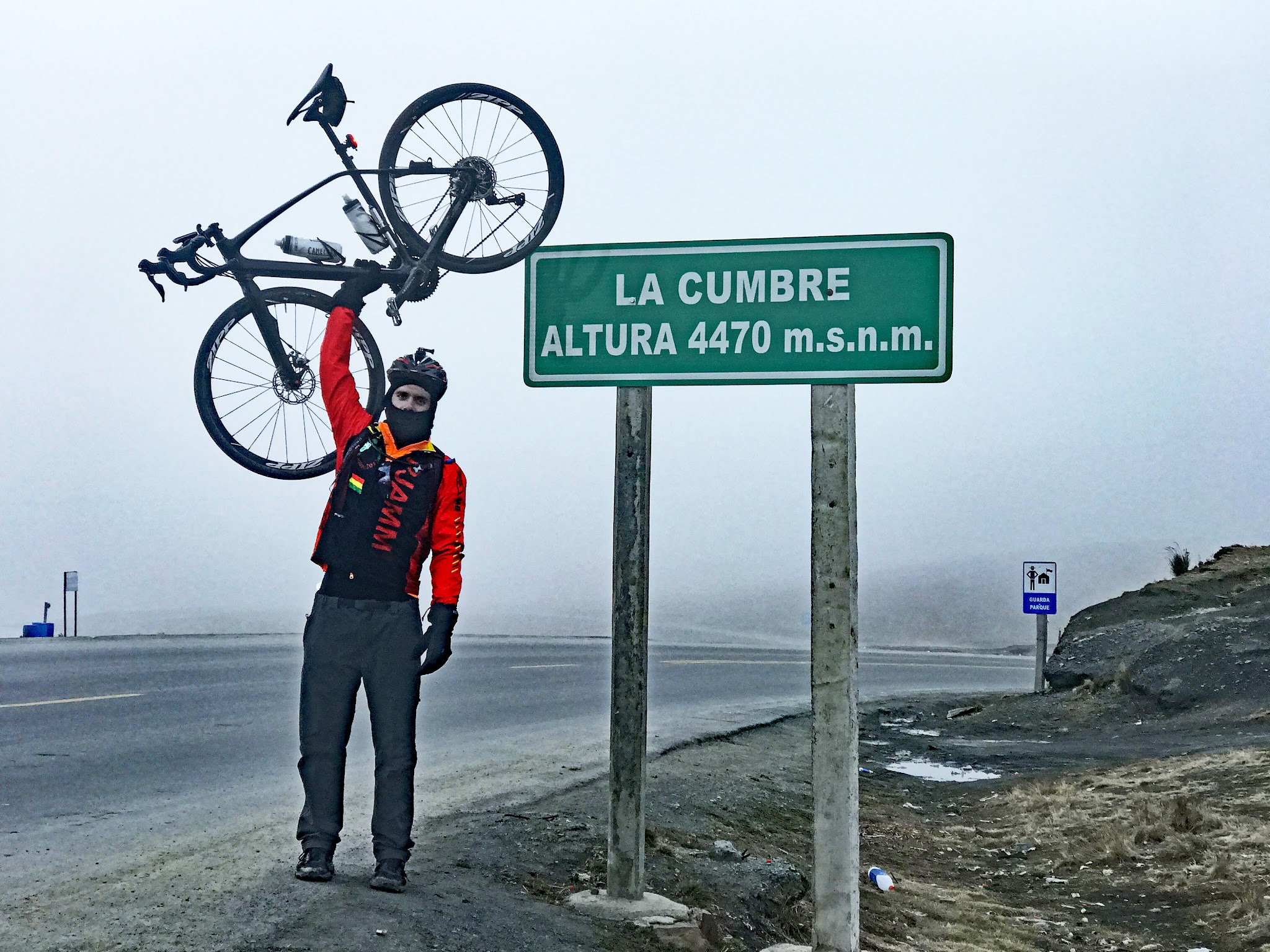 Cyclist raising bike at La Cumbre Peak summit and elevation sign