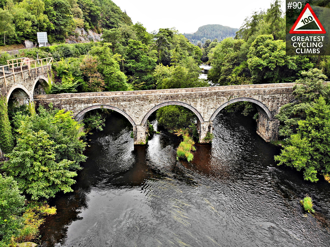 Climbing Horseshoe Pass by Bike - aerial view of Llangollen Bridge, old stone bridge with 3 arches, river passing through, dense foliage in and around river, Simon Warren's 100 Greatest Cycling Climbs #90 Logo