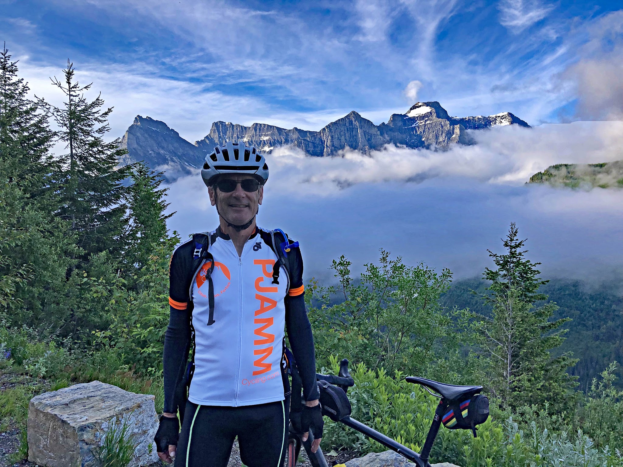 Cycling Logan Pass West, Going-to-the-Sun Road, Glacier National Park - cyclist Bruce Hamilton wears PJAMM Cycling Jersey and stands along low stone retaining wall on Logan Pass West, sharp mountains in background, fog covering much of center-ground