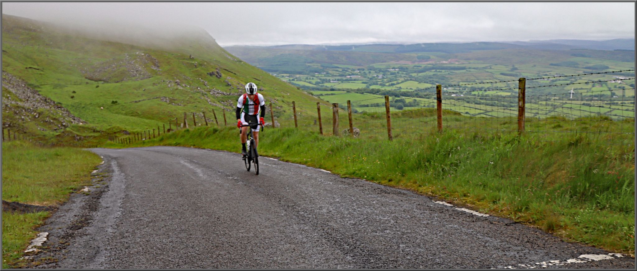 PJAMM Cyclist rides up one-lane country roadway surrounded by green hillsides and pastures