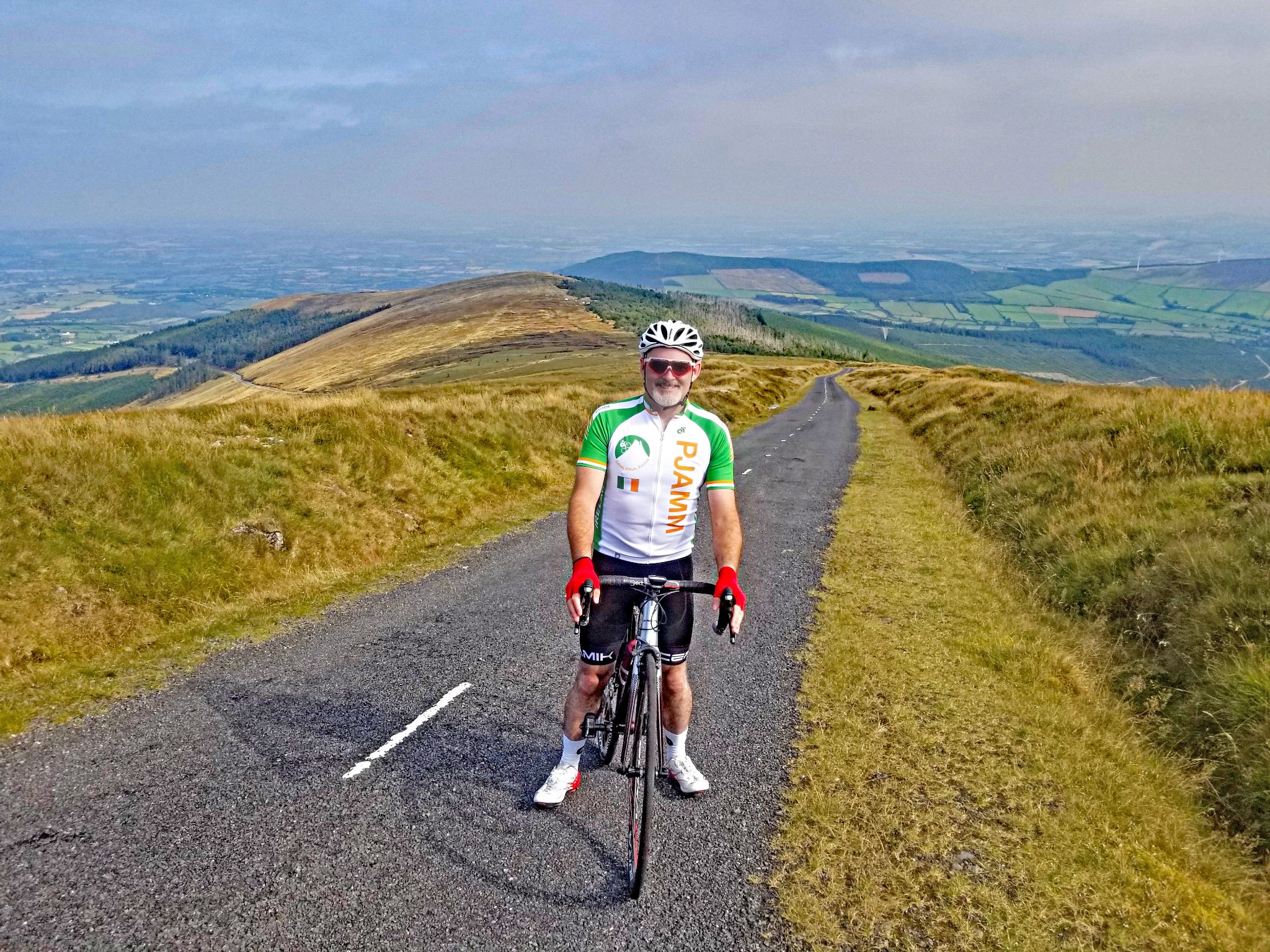 PJAMM Cyclist Stephen Rochford stands with bike in center of narrow two-lane roadway in Irish countryside, rolling green hills behind