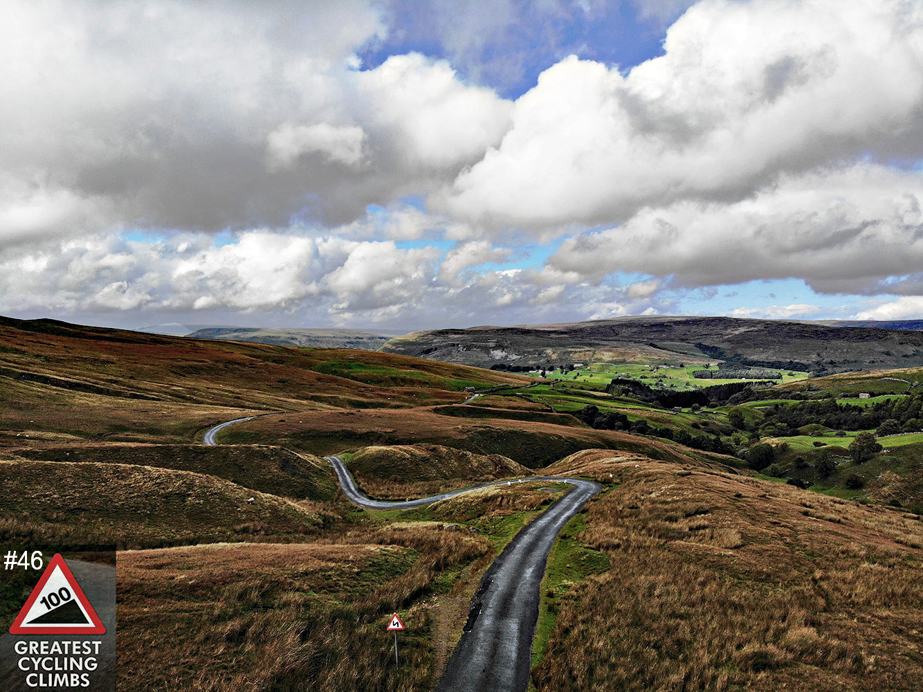 Cycling Oxnop Scar - photo looking down over rolling hills at a narrow roadway winding through the hills, pastureland in background, blue sky with white fluffy clouds, Simon Warren's 100 Greatest Cycling Climbs #46 logo in corner
