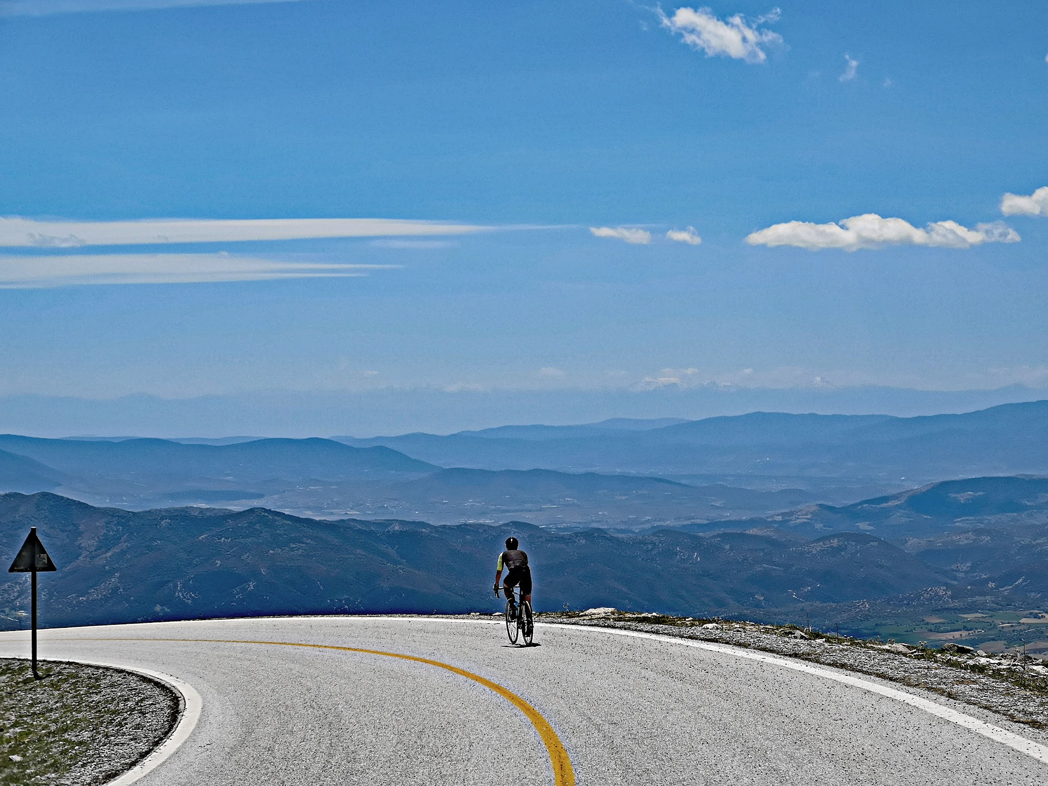 Cycling Mount Olympus, Greece - cyclist rides on curve in two-lane roadway on mountainside, mountains can be seen layering the horizon, white clouds in blue sky