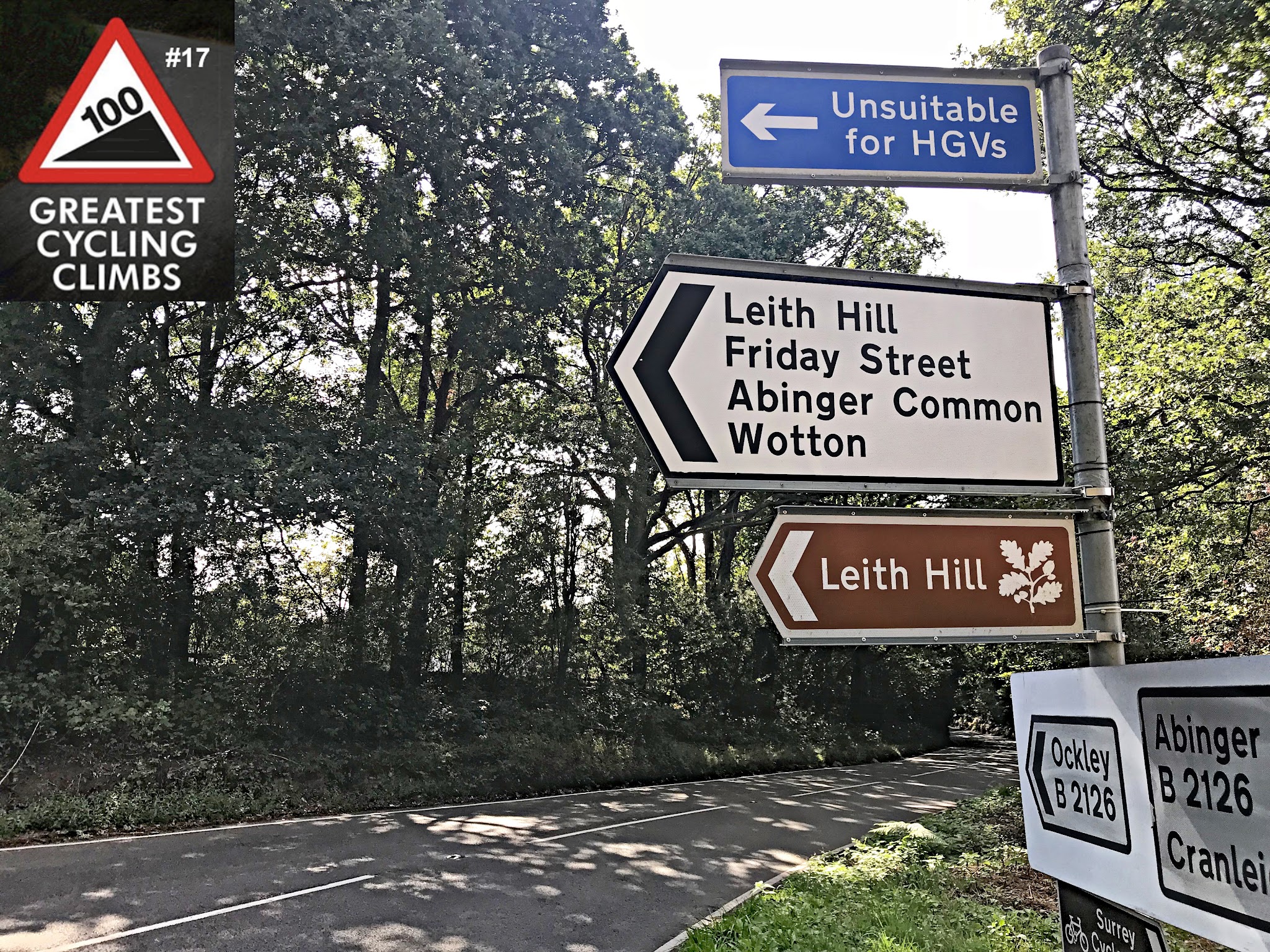 Cycling Leith Hill, England - road signs along two-lane roadway with dense tree lining, signs for Leith Hill, Friday Street, Abinger Common, and Wotton; Simon Warren's 100 Greatest Cycling Climbs #17 logo in corner