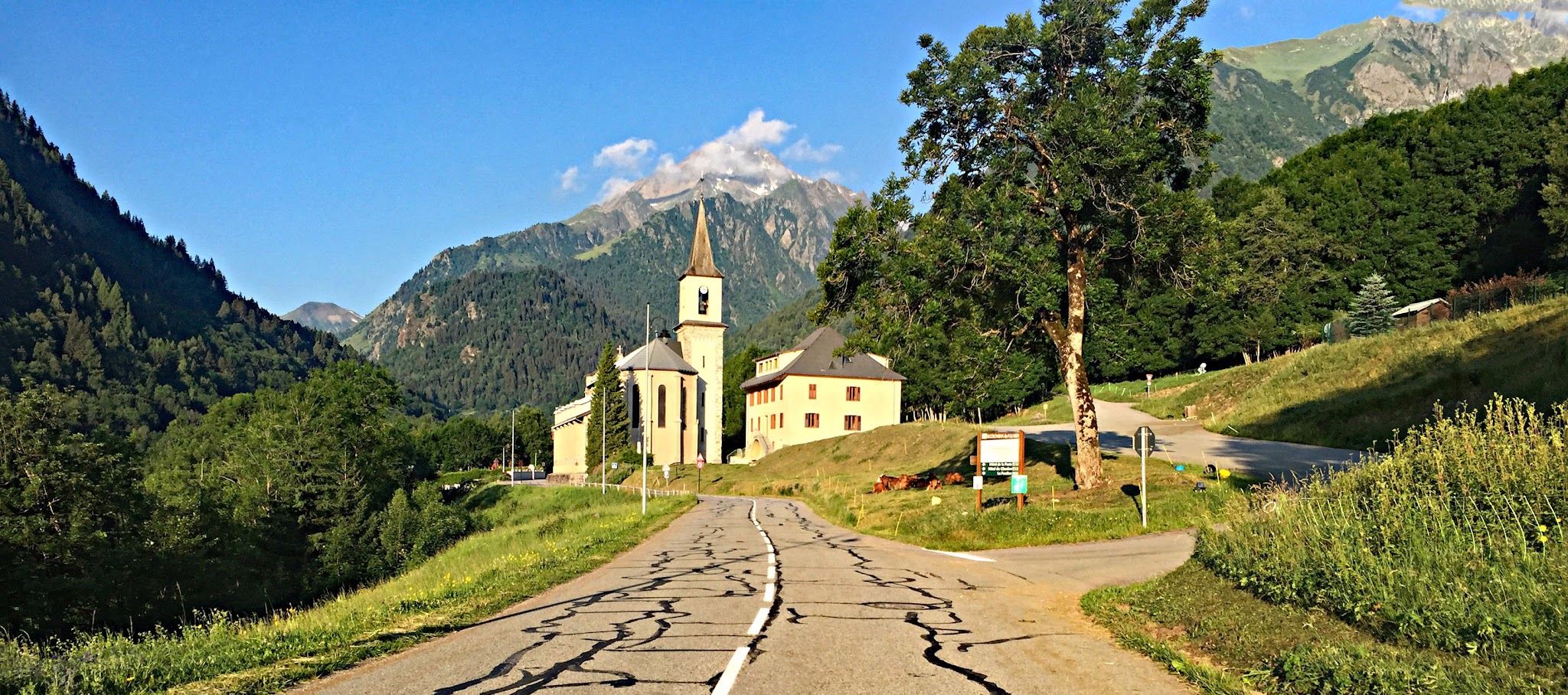 roadway approaches large white church and steeple with mighty French mountains in distance behind church