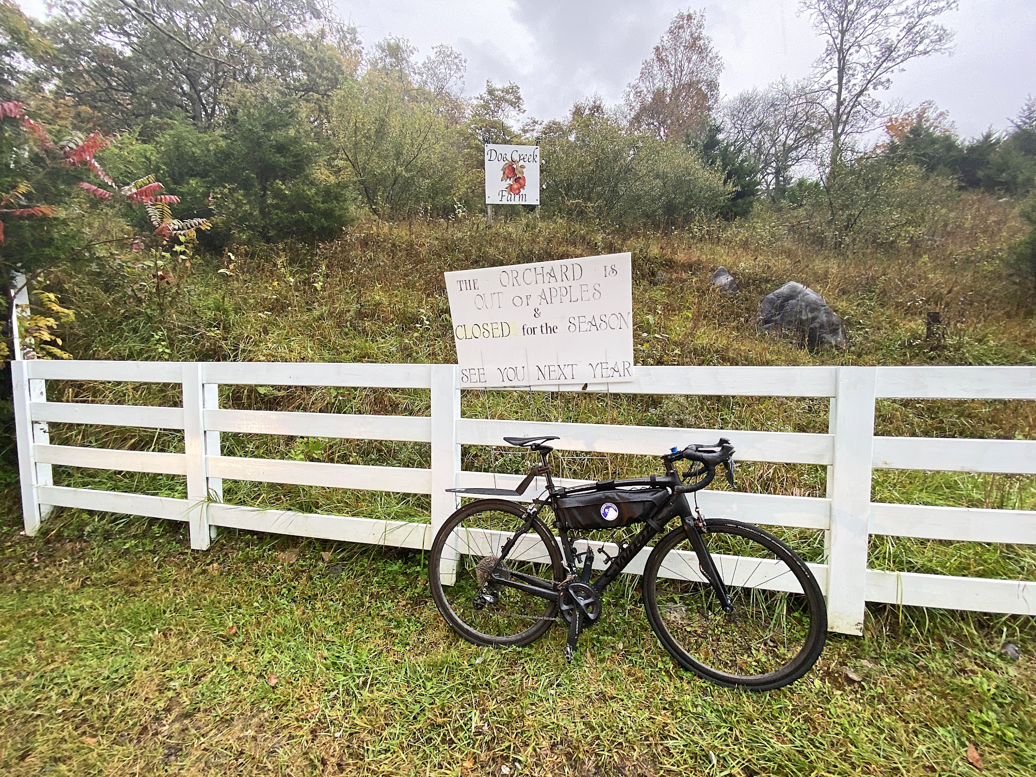 Bike parked against white fence at sign for Doe Creek Farm, Apple orchards