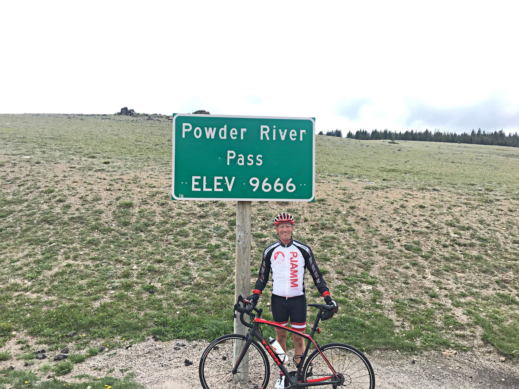 Cycling Powder River, Wyoming - cyclist stands with bike, smiling next to sign for Powder River Pass, Elevation 9666