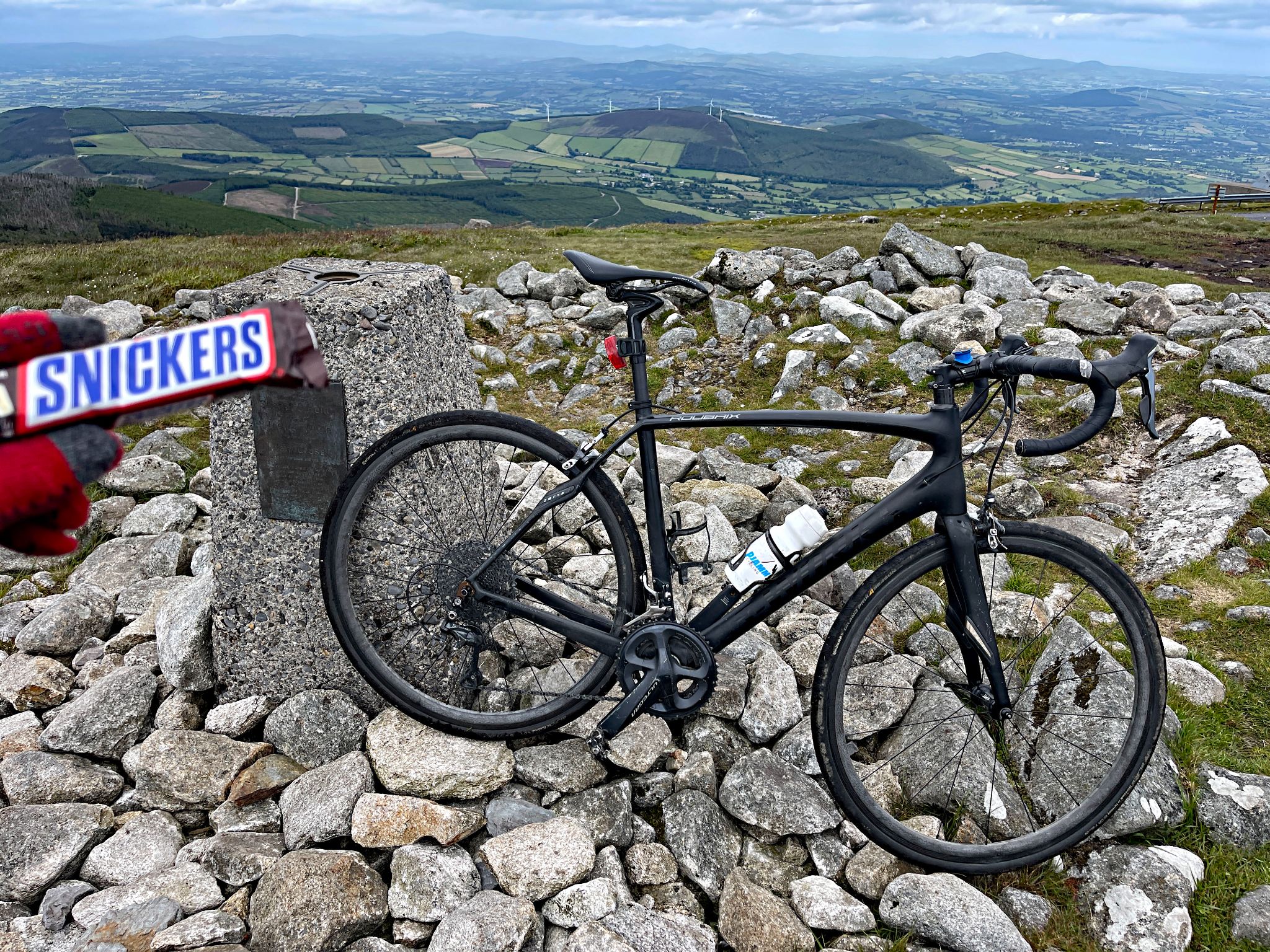 bike parked in stones at climb finish, PJAMM Cyclist holds up snickers bar