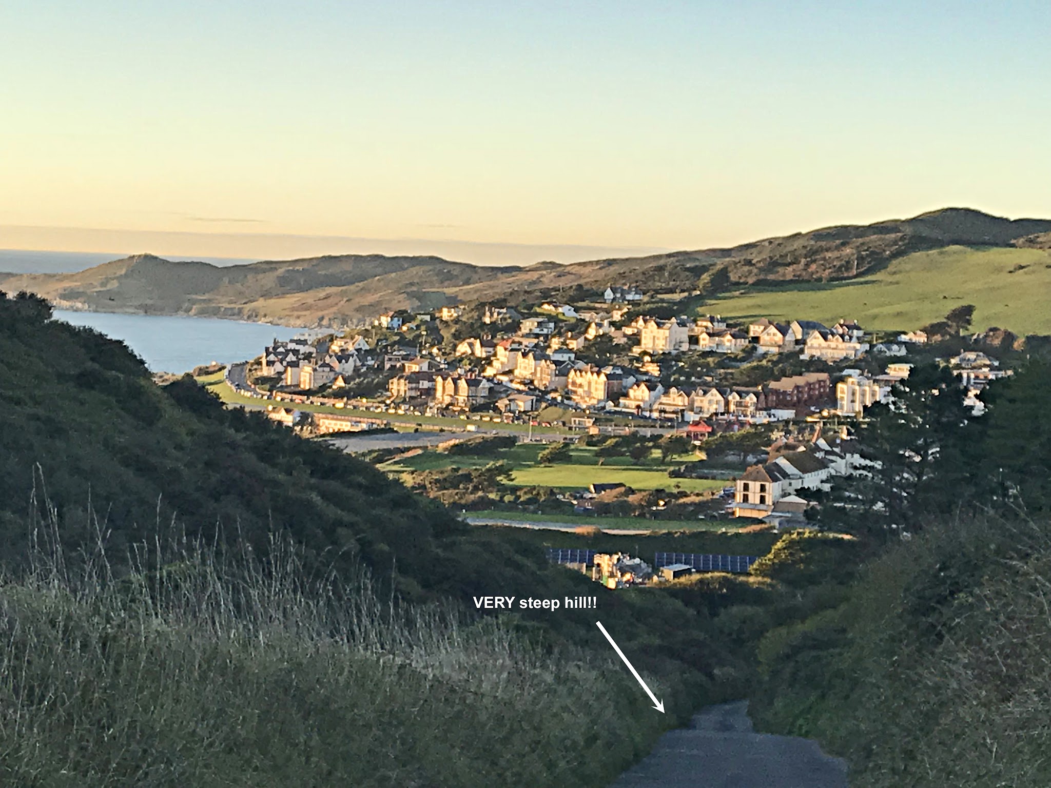 Cycling Challacombe Hill - very steep, very narrow roadway lined with tall grasses, roadway is leading up from a costal village dotted with white buildings and homes, sunset over water in background