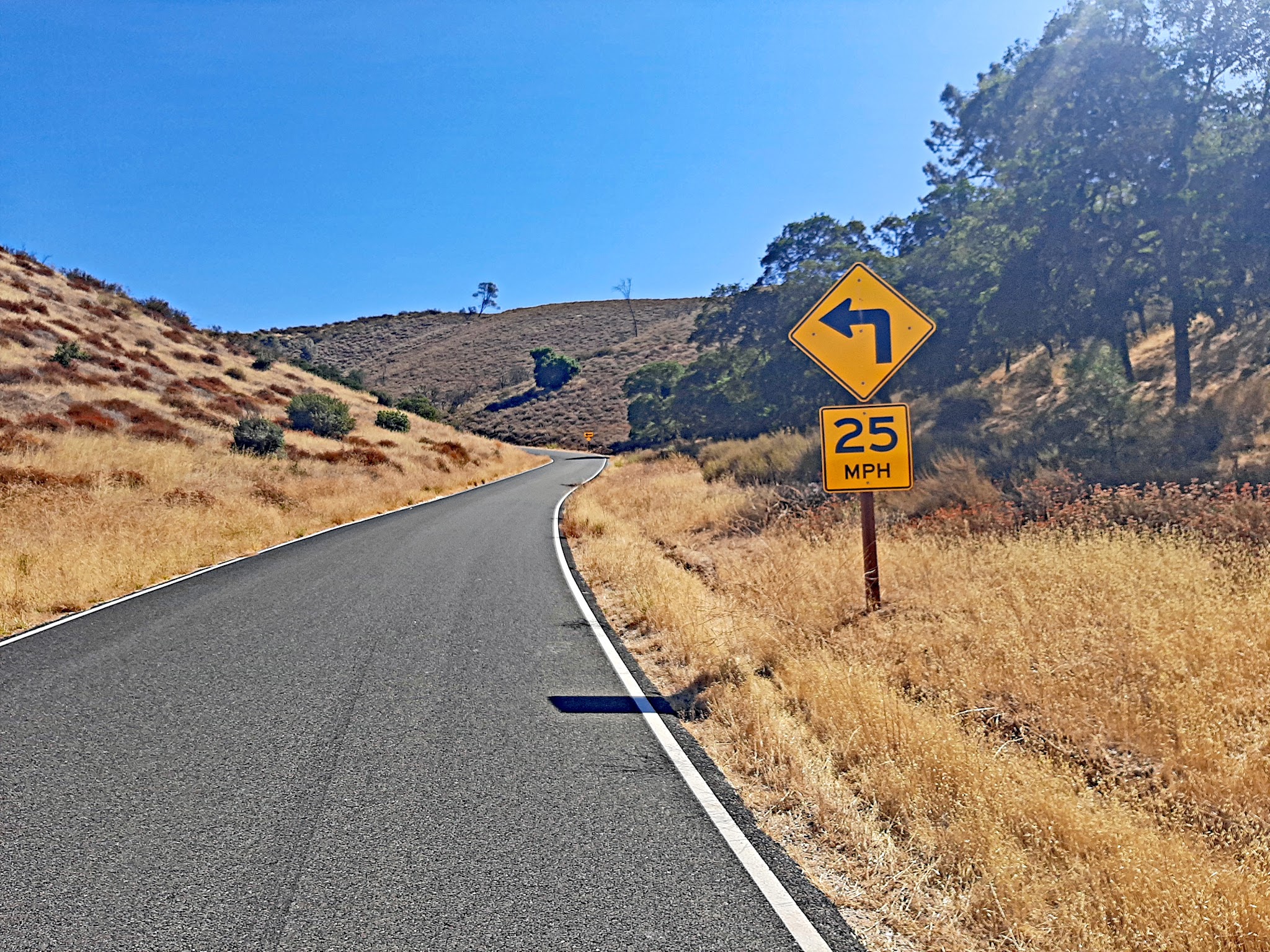 Cycling Pinnacles National Park, California - one-lane road winding through hillsides covered in brown grass and scrub brush, road sign for 25 MPH on turn
