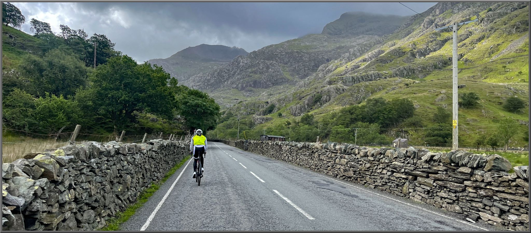 PJAMM Cyclist rides on two-lane road bordered by stone walls and surrounded by green hillsides