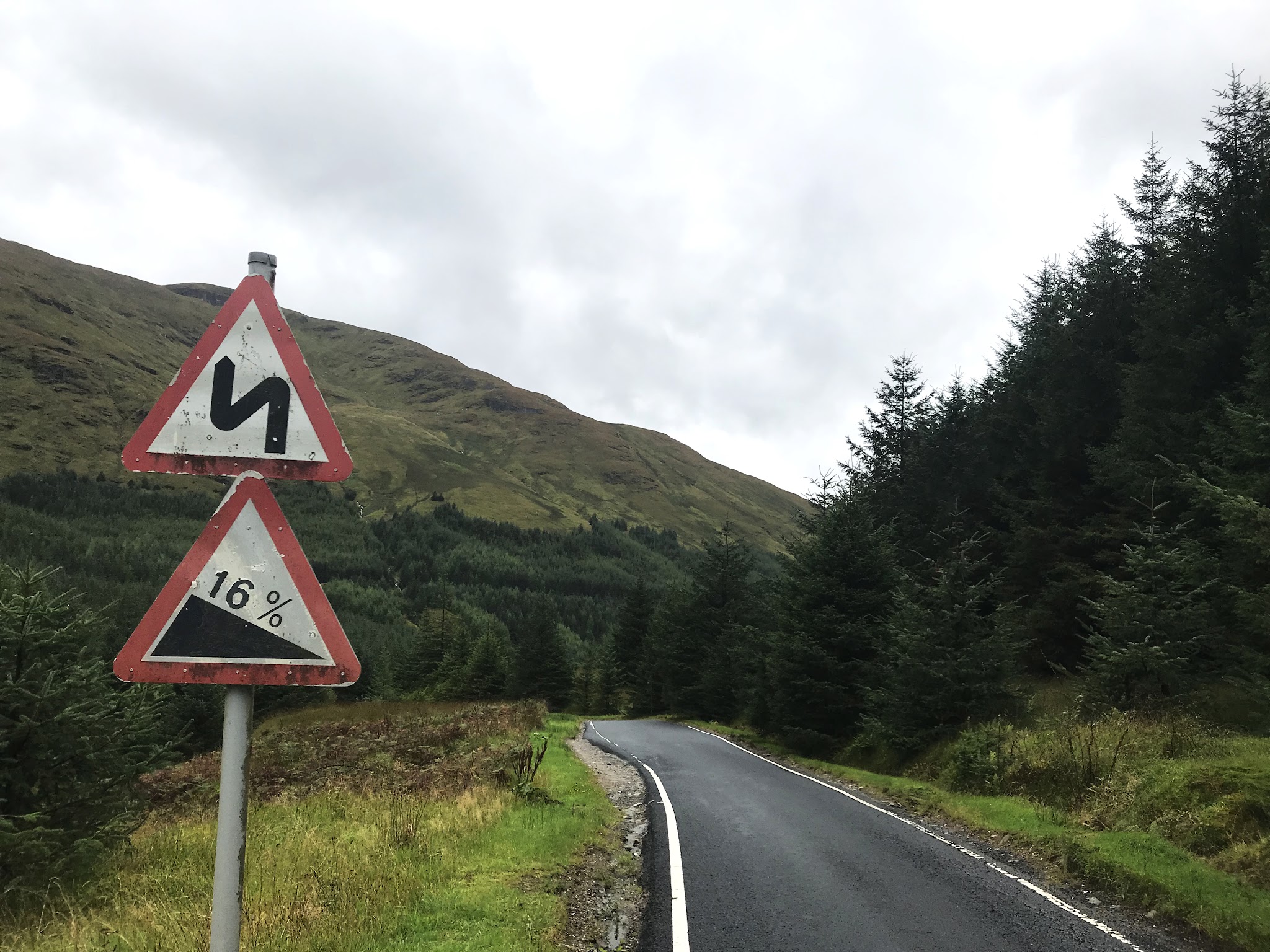 Cycling Rest and Be Thankful, Scotland - straight portion of one-lane road next to road sign noting 16% grades and winding road, lush greenery and pine trees along hillside, cloudy sky