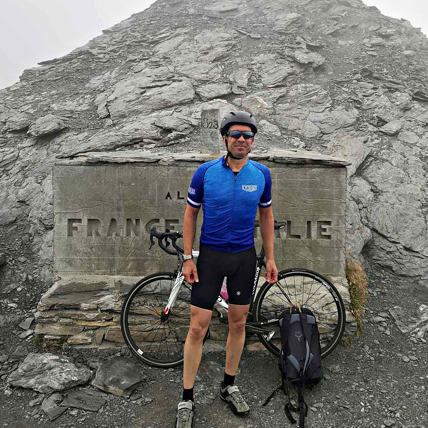 Cycling Colle dell’Agnello, France and Italy - PJAMM Cyclist stands with bike in front of stone sign for border of France and Italy