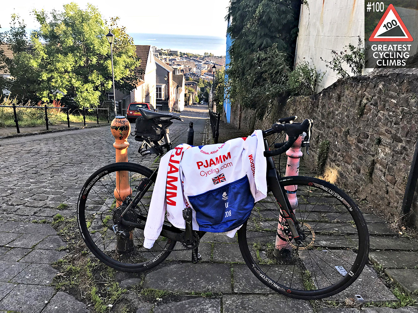 Cycling Constitution Hill, Wales - bike with PJAMM Cycling jersey draped over it, parked at climb top on cobblestone road, Simon Warren's Greatest Cycling Climbs #100 logo in corner