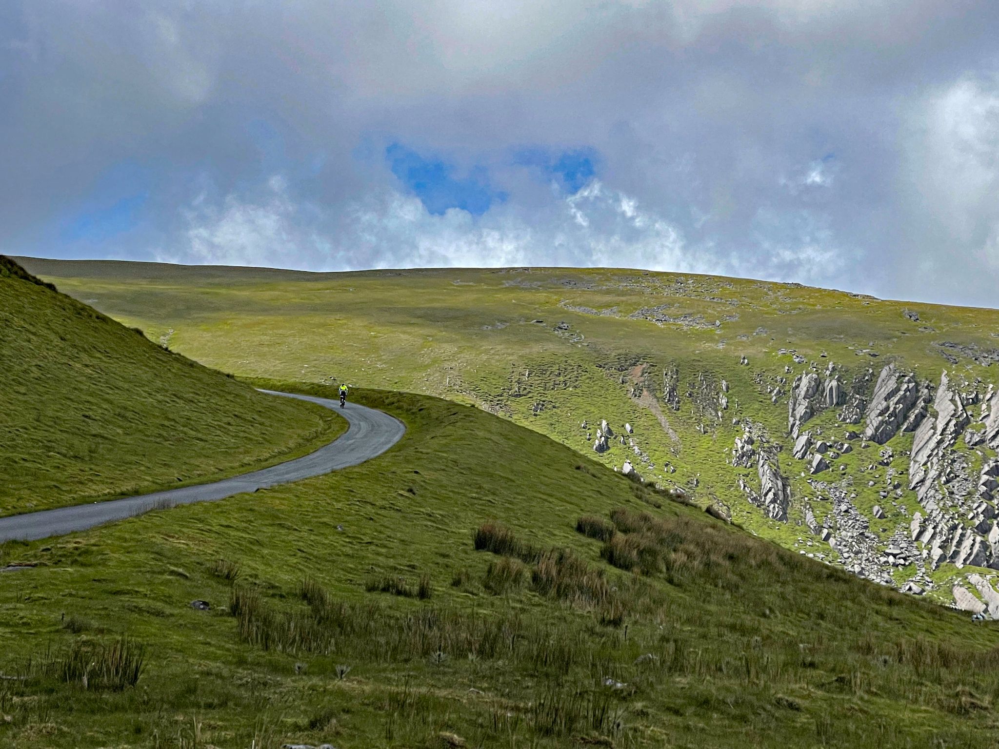 PJAMM Cyclist rides on one-way country road surrounded by lush green hillsides and rocks