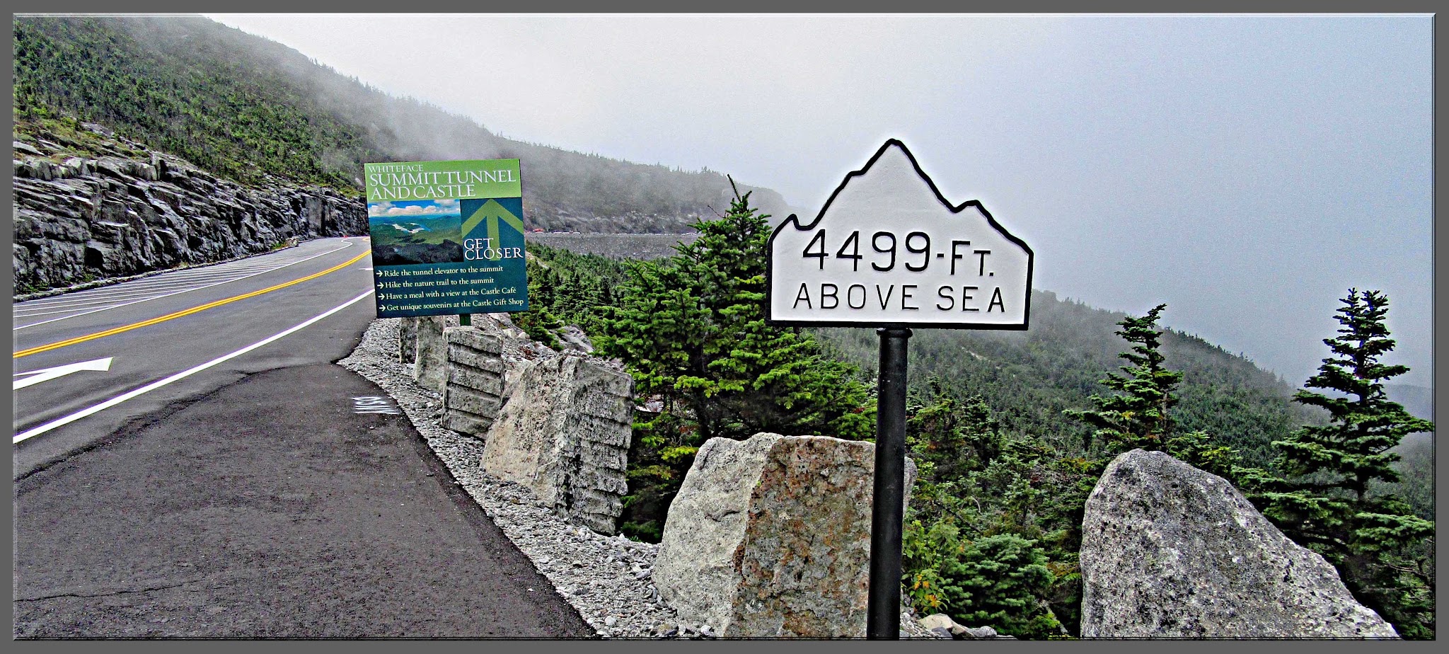 Cycling Whiteface Mountain - Mountain-shaped road sign noting 4499 feet above sea level, side of road along the mountainside with green trees peeking out through a foggy background