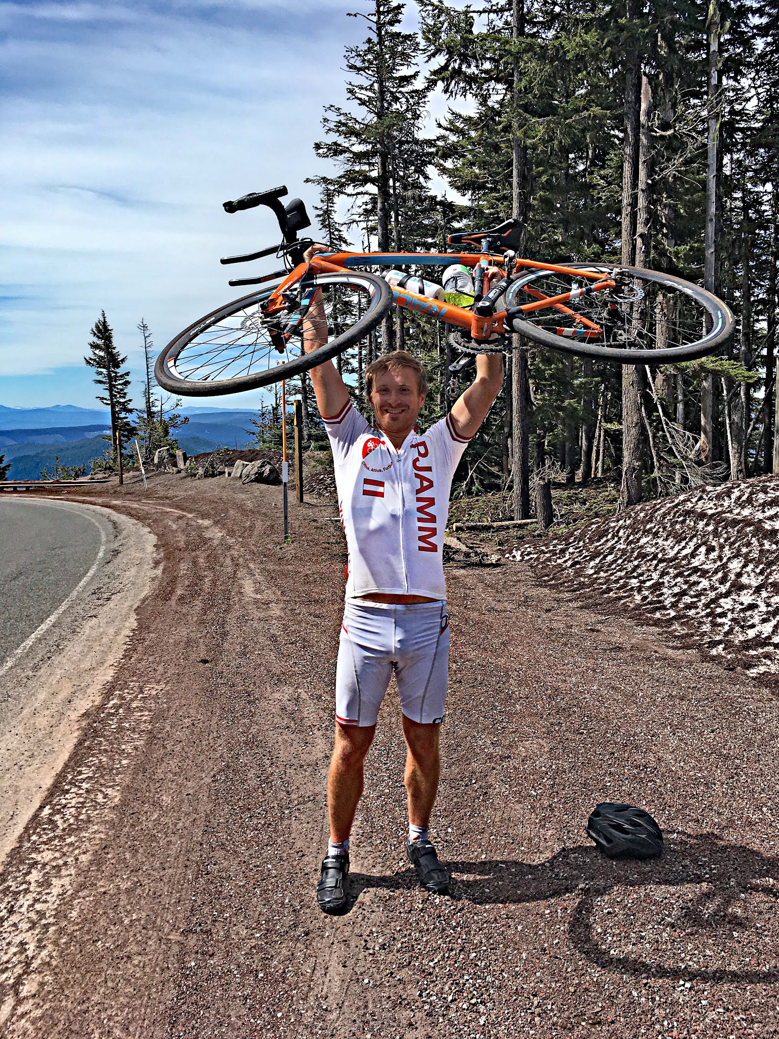 PJAMM Cyclist Garrett Simpson stands at summit holding his bike in the air