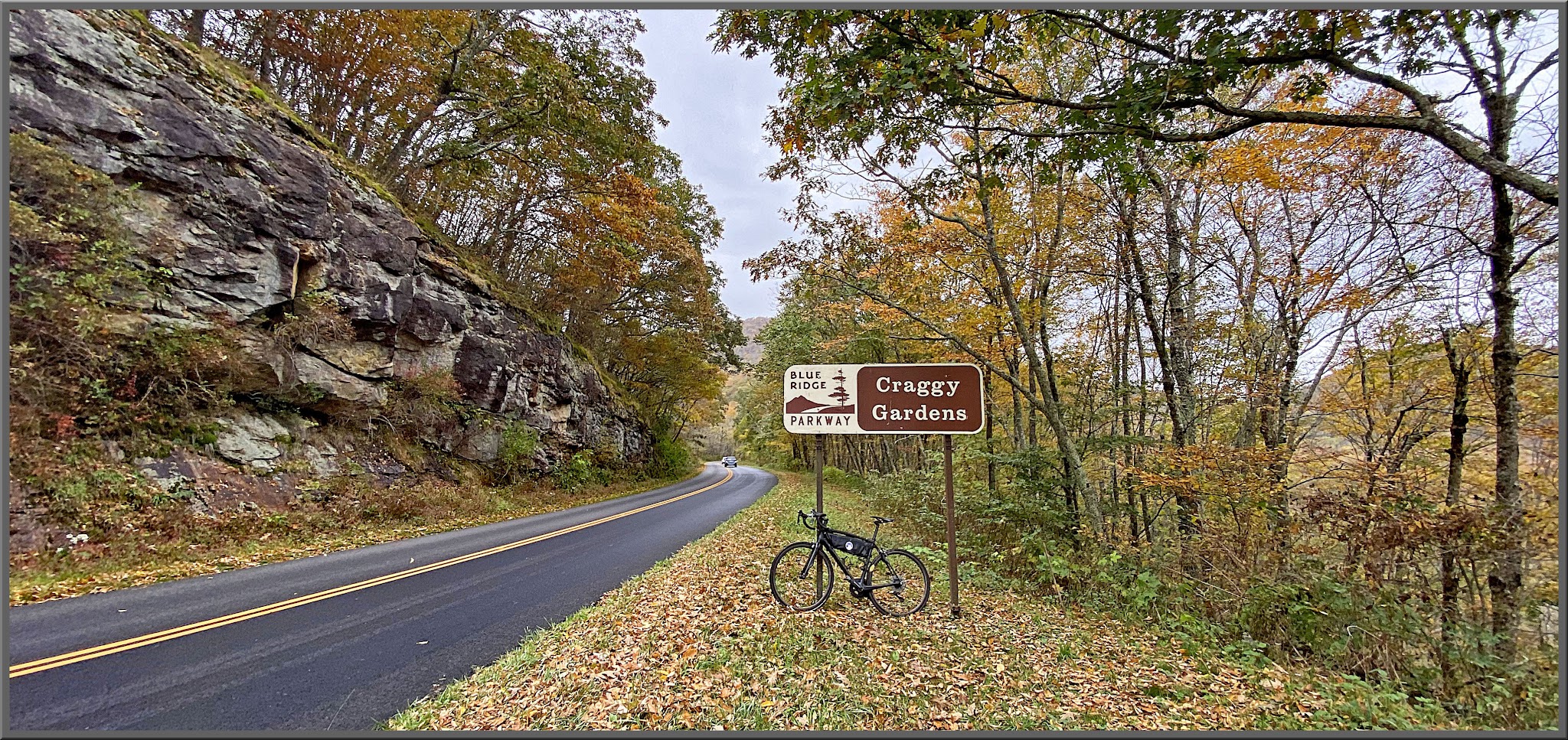Cycling Blue Ridge Parkway to Craggy Gardens - bike parked next to BRP sign for Craggy Gardens, across two-lane roadway from tall stone wall