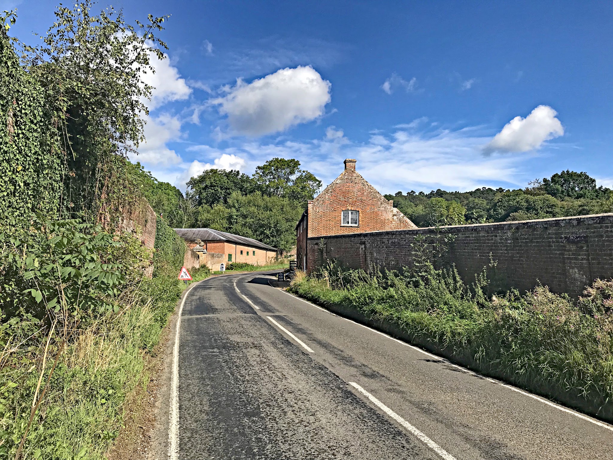 Cycling Leith Hill, England - two-lane road stretching uphill, brick retaining wall on side of road, brick building, vegetation on side of road, blue sky with white clouds