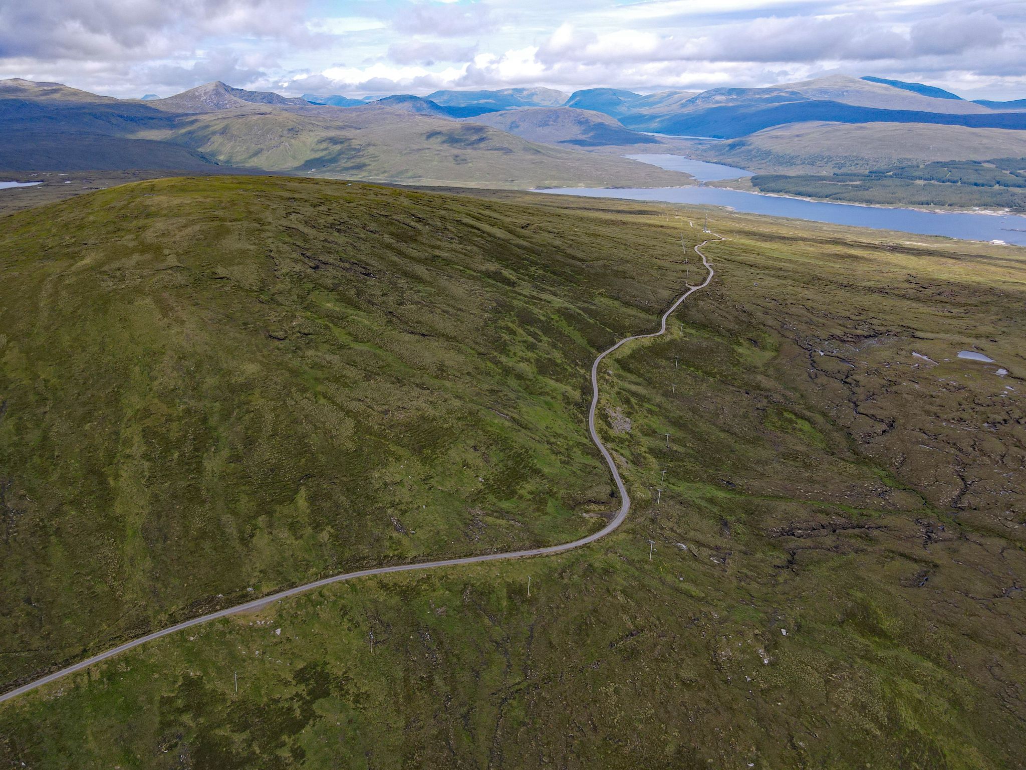 aerial drone view shows panoramic view of roadway leaving Loch Shin behind and traveling up mountainside to Scottish Highlands