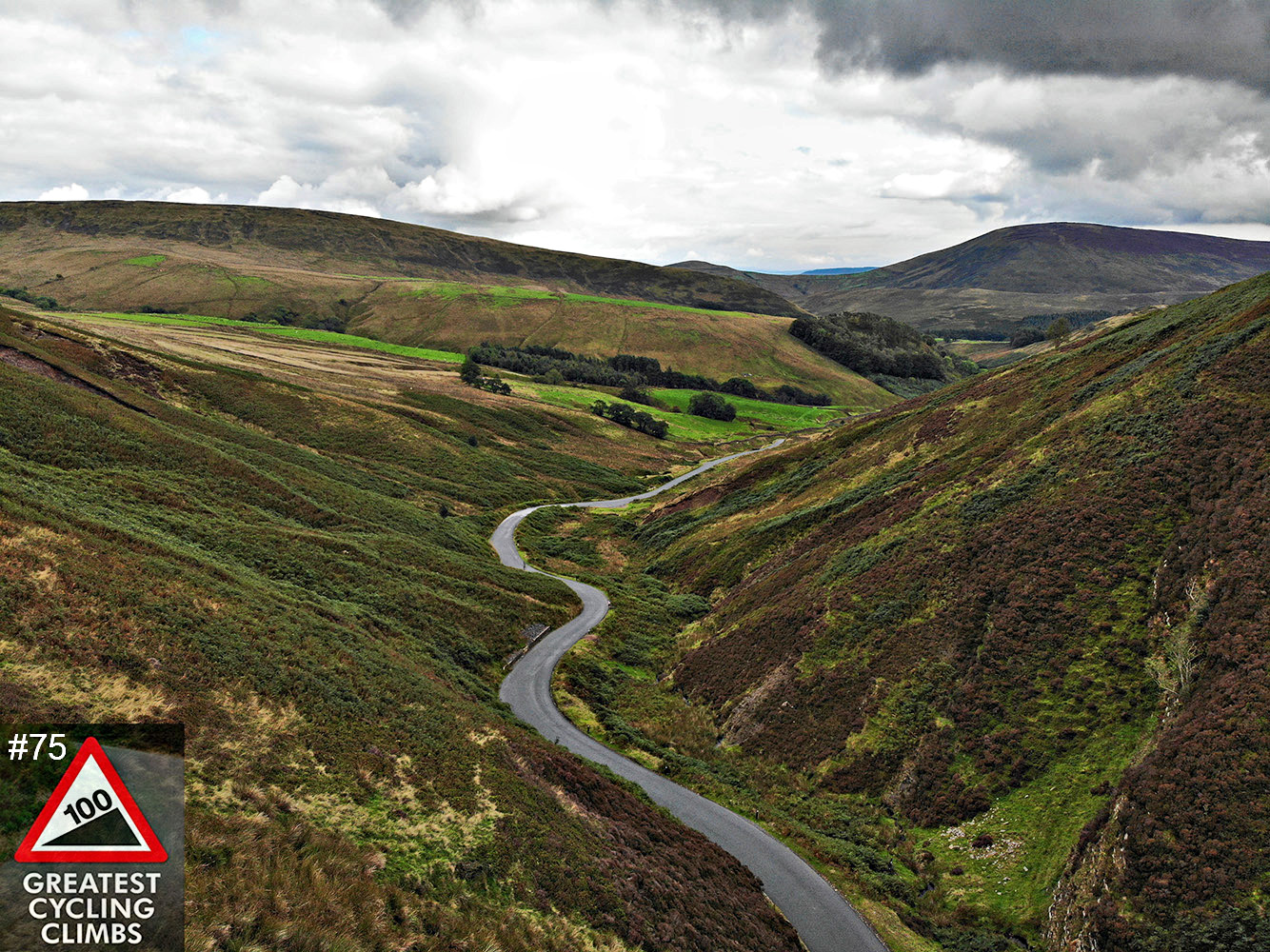 Cycling Trough of Bowland, Britain - aerial view of winding road passing through a valley between two brown and green hillsides, cloudy sky, 100 Greatest Cycling Climbs #75 logo in corner