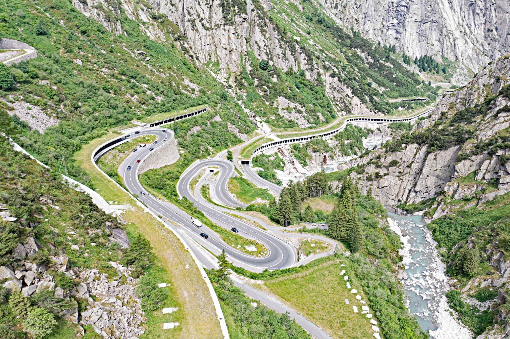aerial drone view of road snaking up mountainside, switchbacks all the way; far beneath is a rushing river