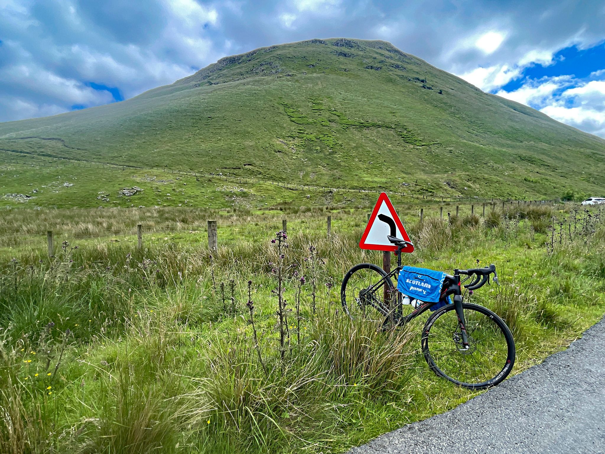 bike parked against road sign warning of curve in road, large green hillside behind