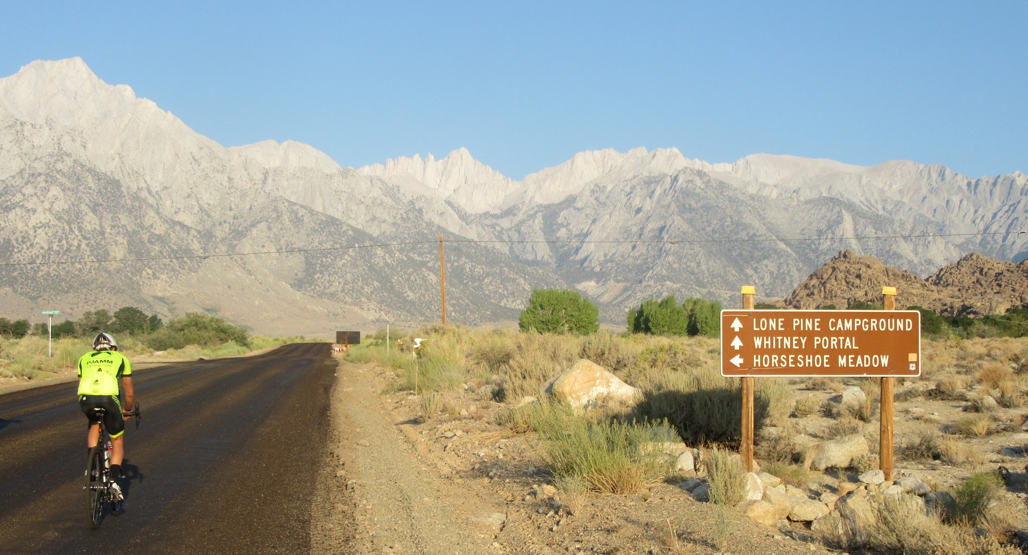 Cyclist on highway roadway with Mt. Whitney's serrated edge peak in the distance, sign for Lone Pine Campground, Whitney Portal, and Horseshoe Meadow
