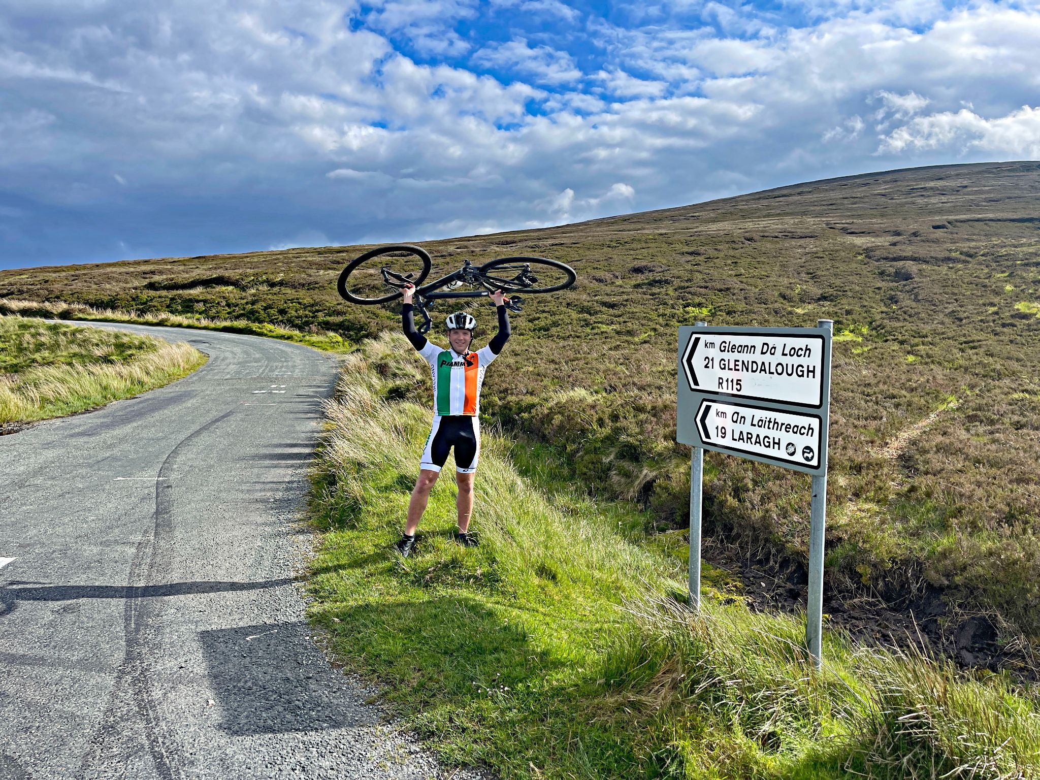 PJAMM Cyclist holds bike above his head at climb finish next to road signs for Glendalough and Laragh