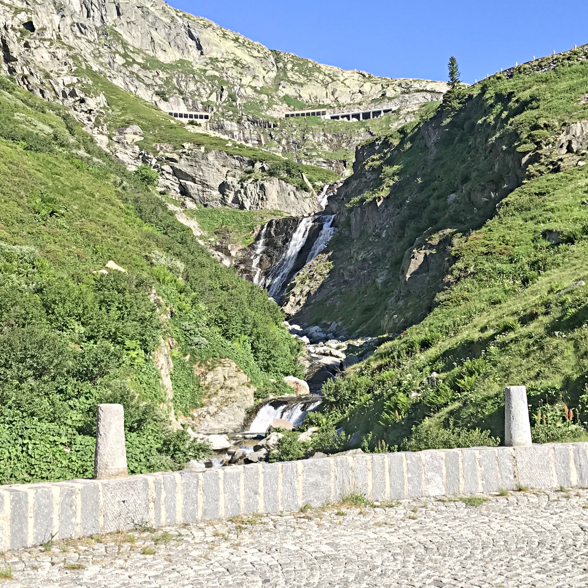 Cycling Gotthard Pass (St. Gotthard Pass), Switzerland - a small waterfall traverses the mountainside next to cobblestone segment of roadway