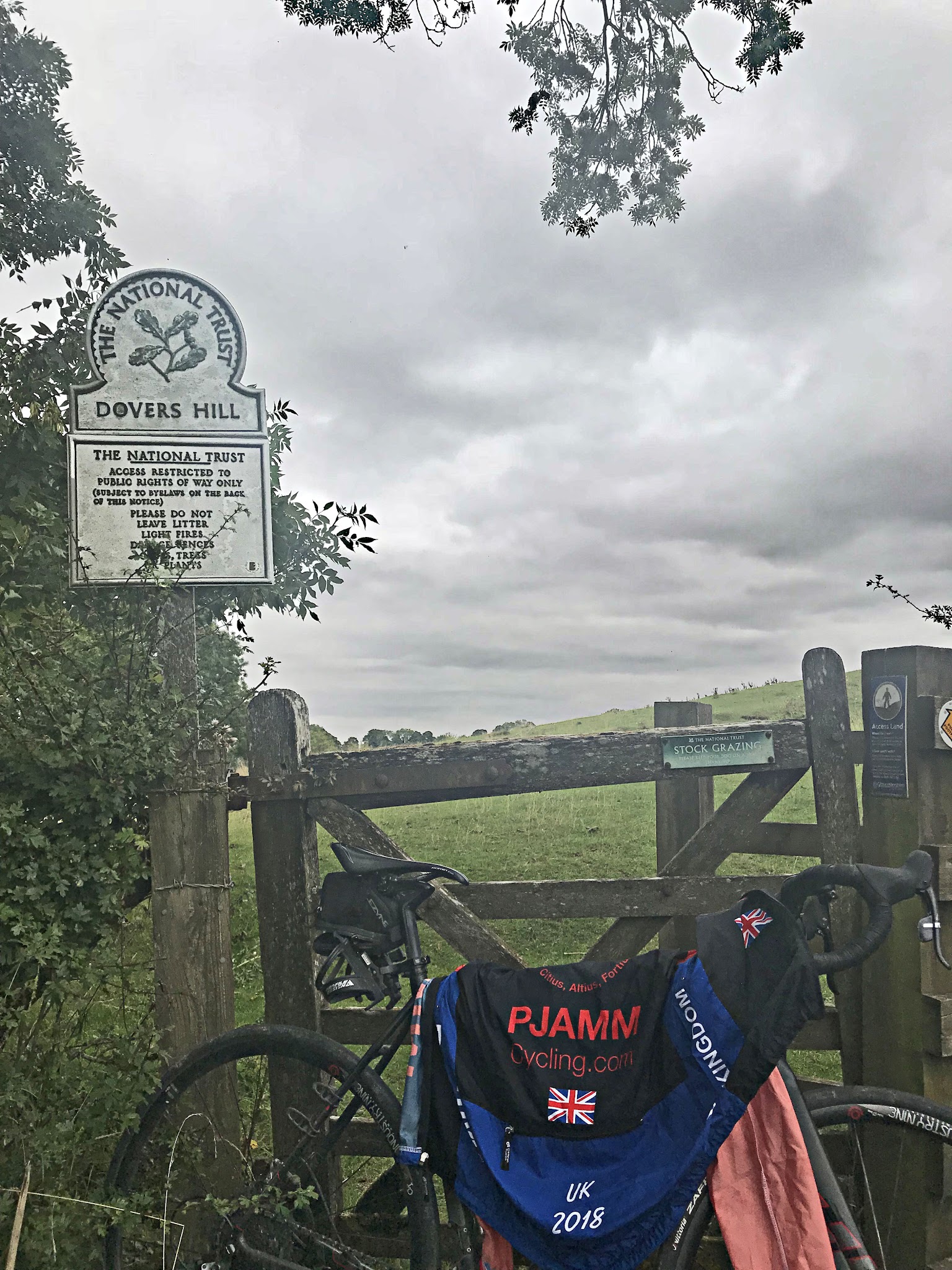 Cycling Dovers Hill - PJAMM Cycling jersey draped over bike leaning against wooden fence along pastureland, The National Trust sign for Dovers Hill