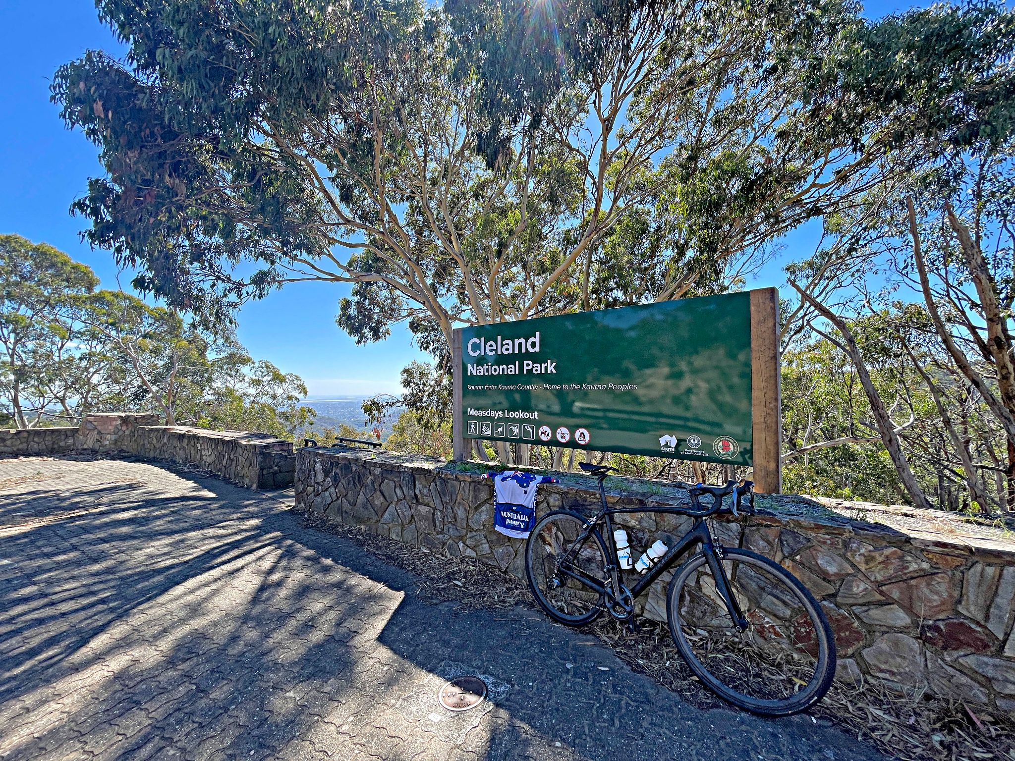 bike and PJAMM Cycling Australia jersey lean against low stone wall in front of Cleland National Park sign