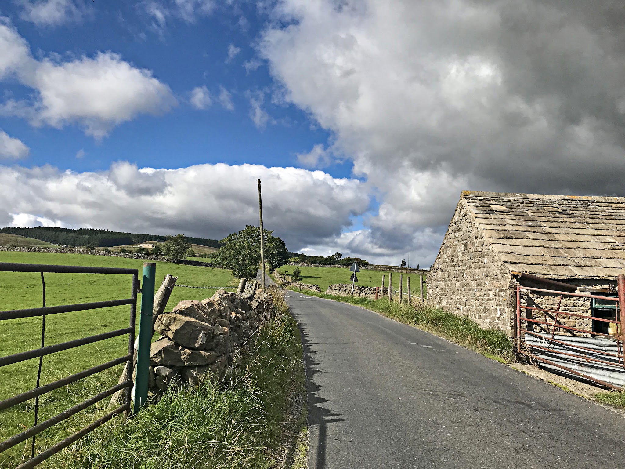 Cycling Chapel Fell - one lane road with pastureland alongside it, low stone fence, old stone building along side of road, blue sky with large white clouds