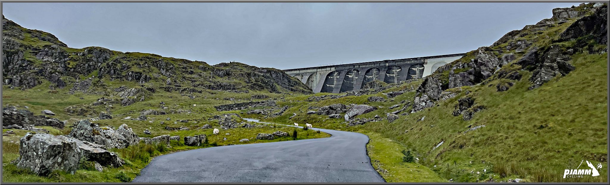 view of one-lane roadway surrounded by green pastureland and rocky hillside, huge dam in middle background