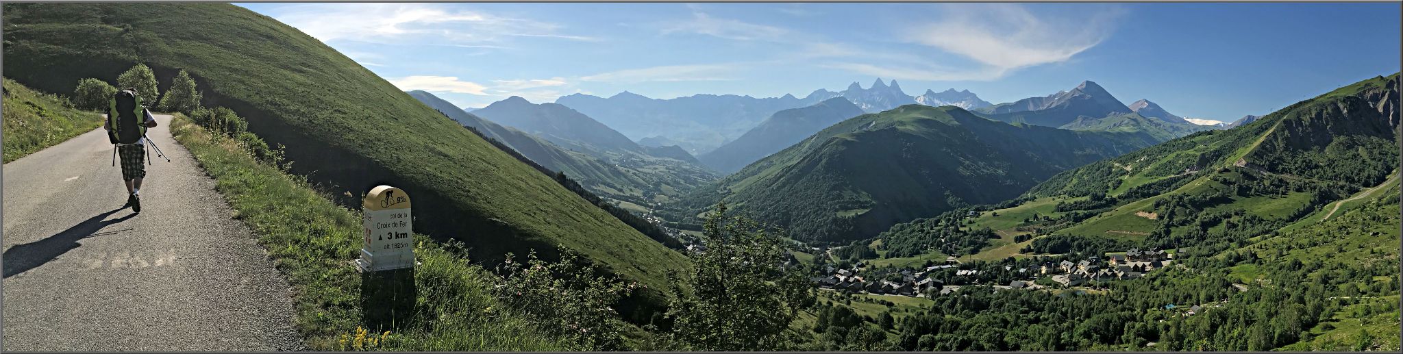 Cycling Col d la Croix de Fer from Saint-Jean-de-Maurienne - panoramic view shows hiker walking on roadway next to KM marker sign, green mountains