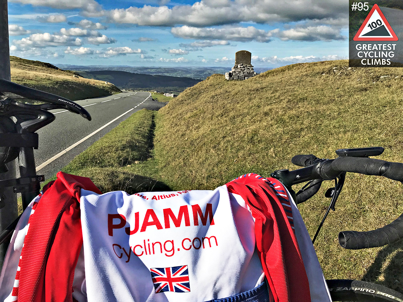 Cycling Black Mountain, Wales - PJAMM Cycling jersey draped over bike parked along side of road near hillside, stone monument in background, blue cloudy skies, Simon Warren's #95 100 Greatest Cycling climbs logo in corner