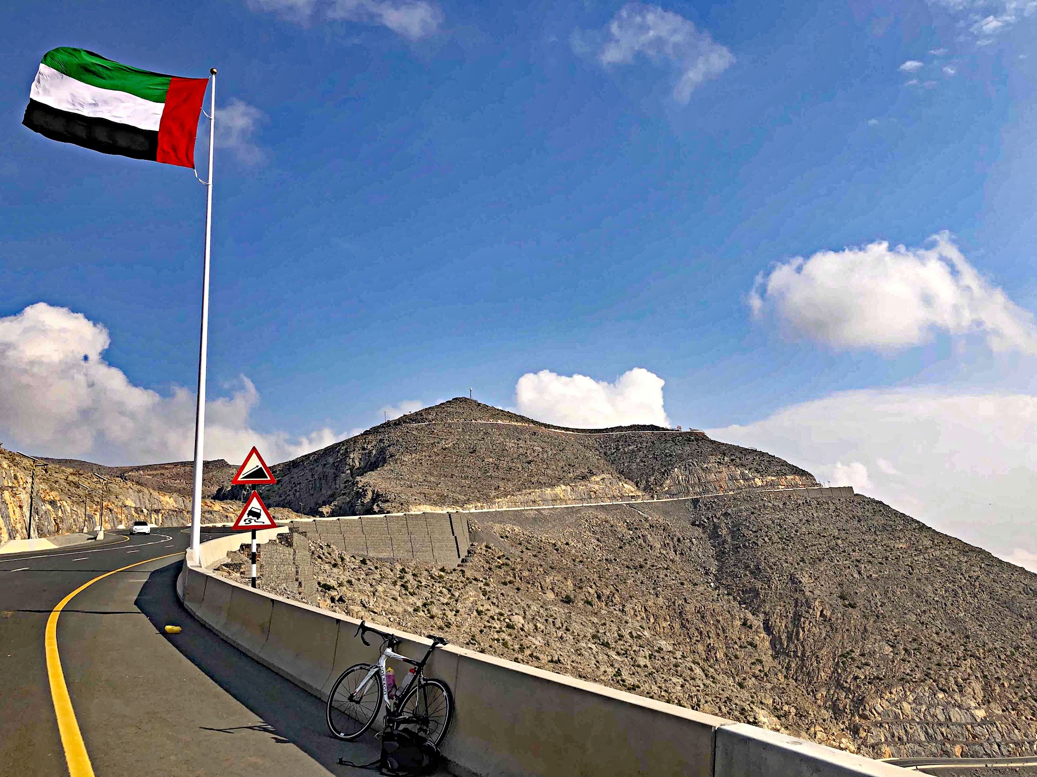 Jebel Jais, Ras Al Khaimah, UAE - bike parked on roadside on side of mountain, flag for the United Arab Emirates waves on roadside next to road signs warning of steep grade