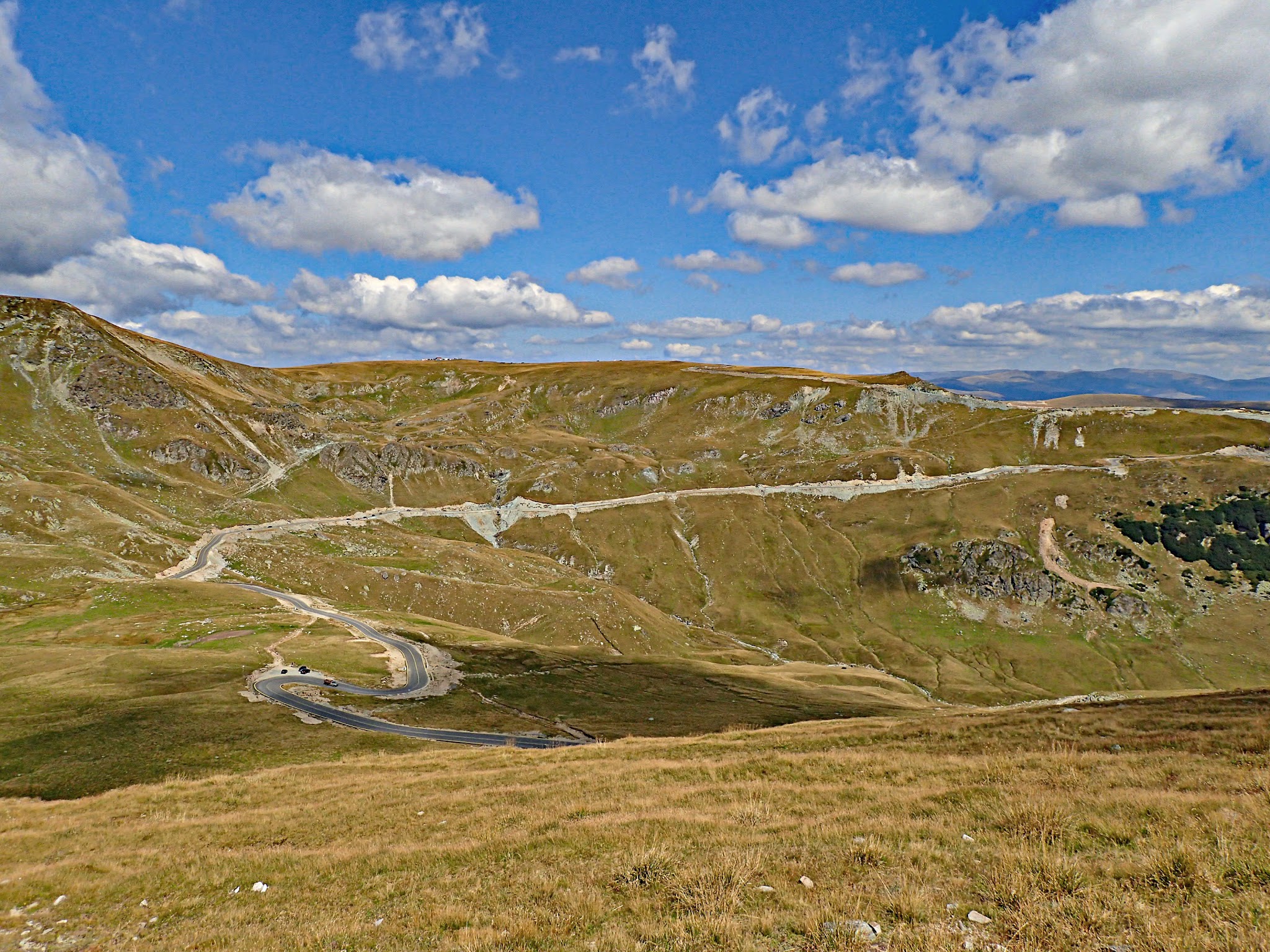 Cycling Transalpina North, Romania - yellow-green grassy hillsides with the Transalpina roadway snaking through, bright blue sky and white clouds