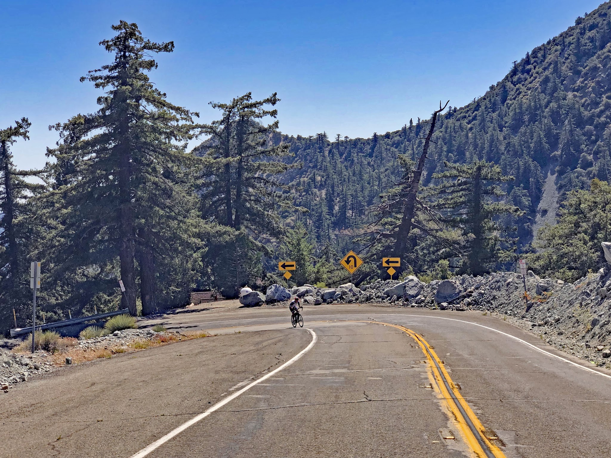 PJAMM Cyclist rides up roadway on Mt. Baldy bike climb, past a hairpin turn in the roadway; alpine setting
