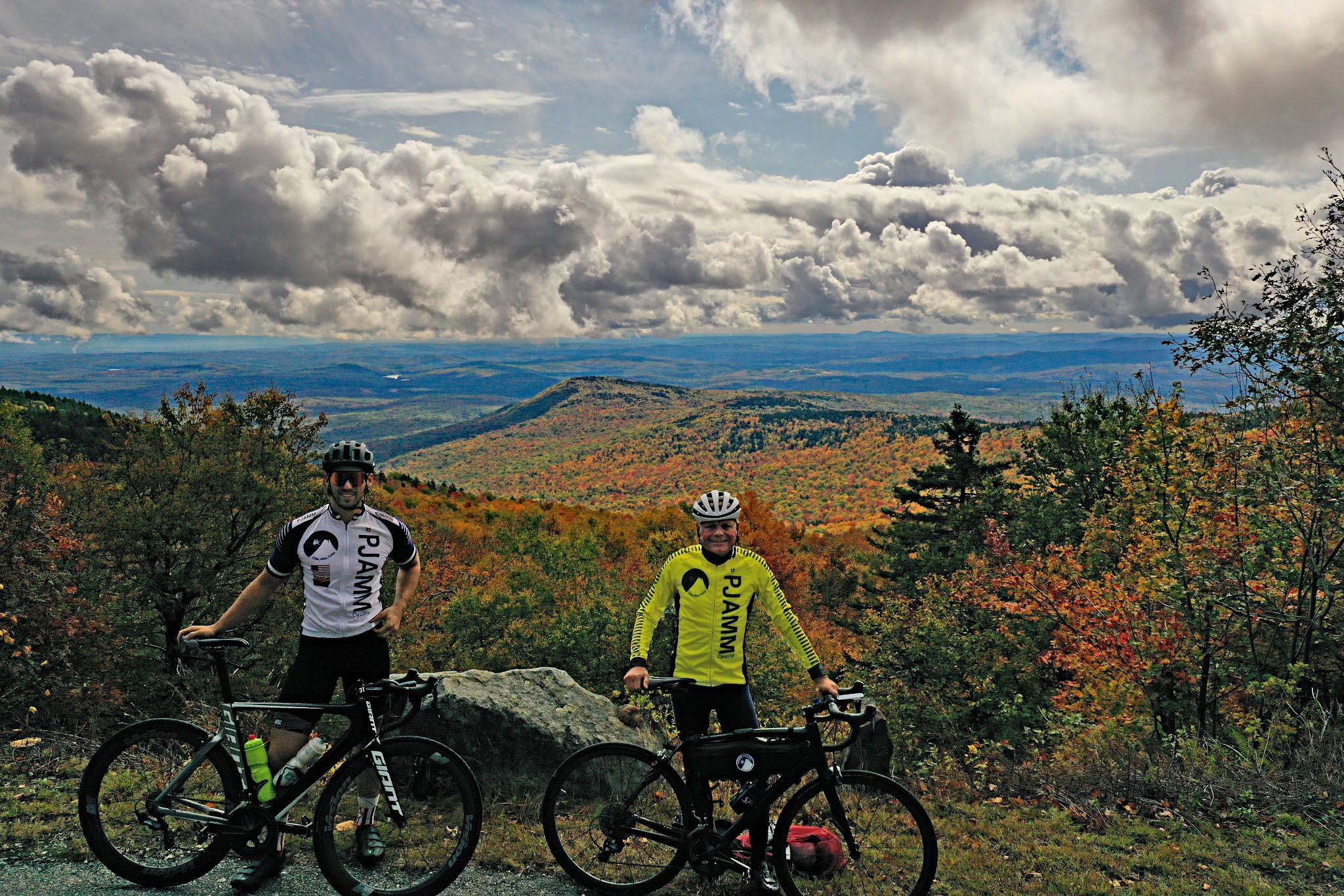 Cycling Kearsarge Mountain Road, New Hampshire - two PJAMM Cyclists stand with bikes atop finish Kearsarge Mountain Road, views behind them are panoramic of dense and bright fall foliage, mountains in background