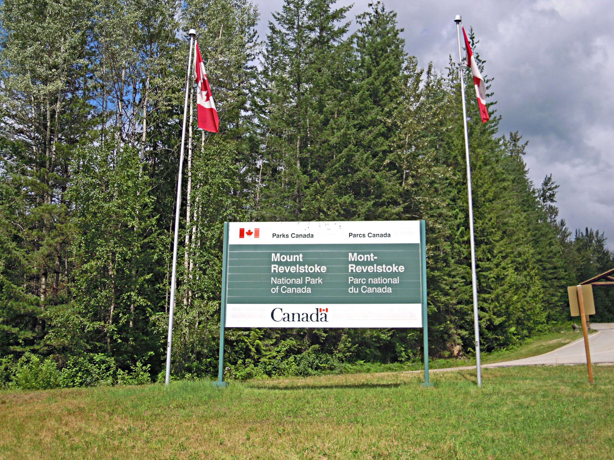 Cycling Mt. Revelstoke, BC, Canada - sign for Mt Revelstoke National Park, two tall Canadian flags on either side, large evergreen trees behind